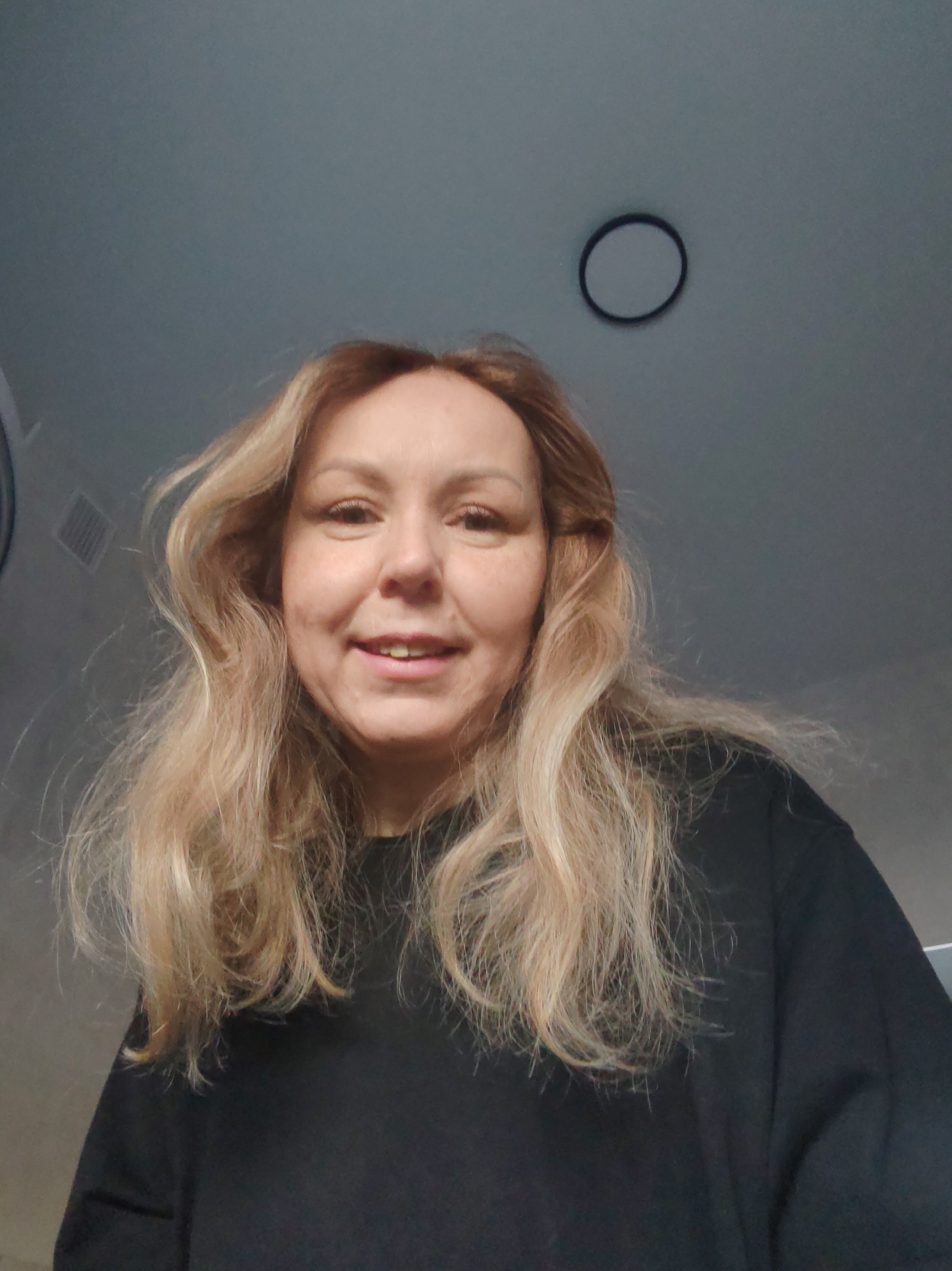A woman with blonde hair and a black shirt is looking directly at the camera. Above her is a white ceiling with a circular light fixture. There is a vent on the wall to the left of her head. I cannot determine if this is in Chaikovsky, Russia.