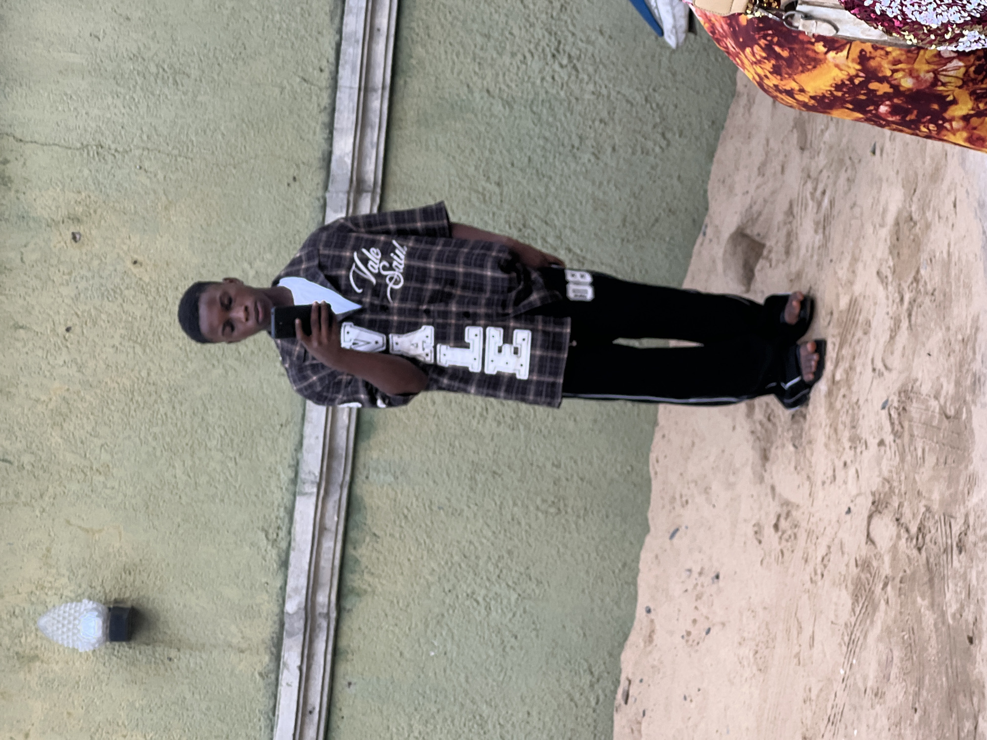 A young man stands on a dirt or sand surface, holding a mobile phone in his right hand, apparently taking a photograph of himself. He wears a plaid shirt with the words "Vale Saint" on it, along with black pants and open-toe sandals. To the left, a green textured wall with a decorative light fixture is visible, marked by vertical trim. To the right, a person partially enters the frame wearing patterned clothing. The exact location within the city of Unknown is not identifiable.