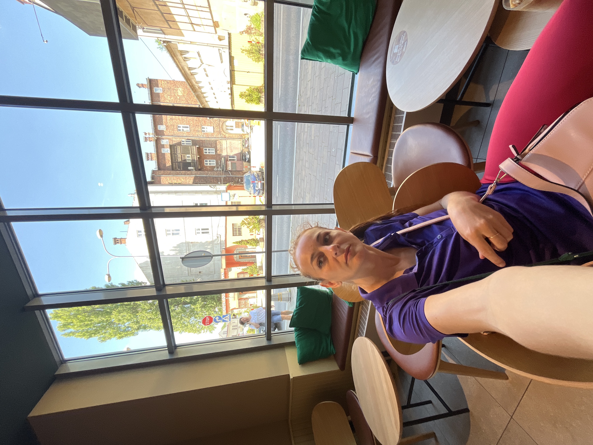 A woman is taking a selfie in a cafe, seated at a table by a large window overlooking a street. She is wearing a purple polo shirt and bright pink leggings, with a light pink handbag slung over her shoulder. In the background, outside the window, a woman in a white t-shirt and blue shorts is walking and talking on her phone. The street scene includes buildings, trees, parked cars, and streetlights. The cafe interior features wooden tables and chairs, and green cushions on a built-in bench.