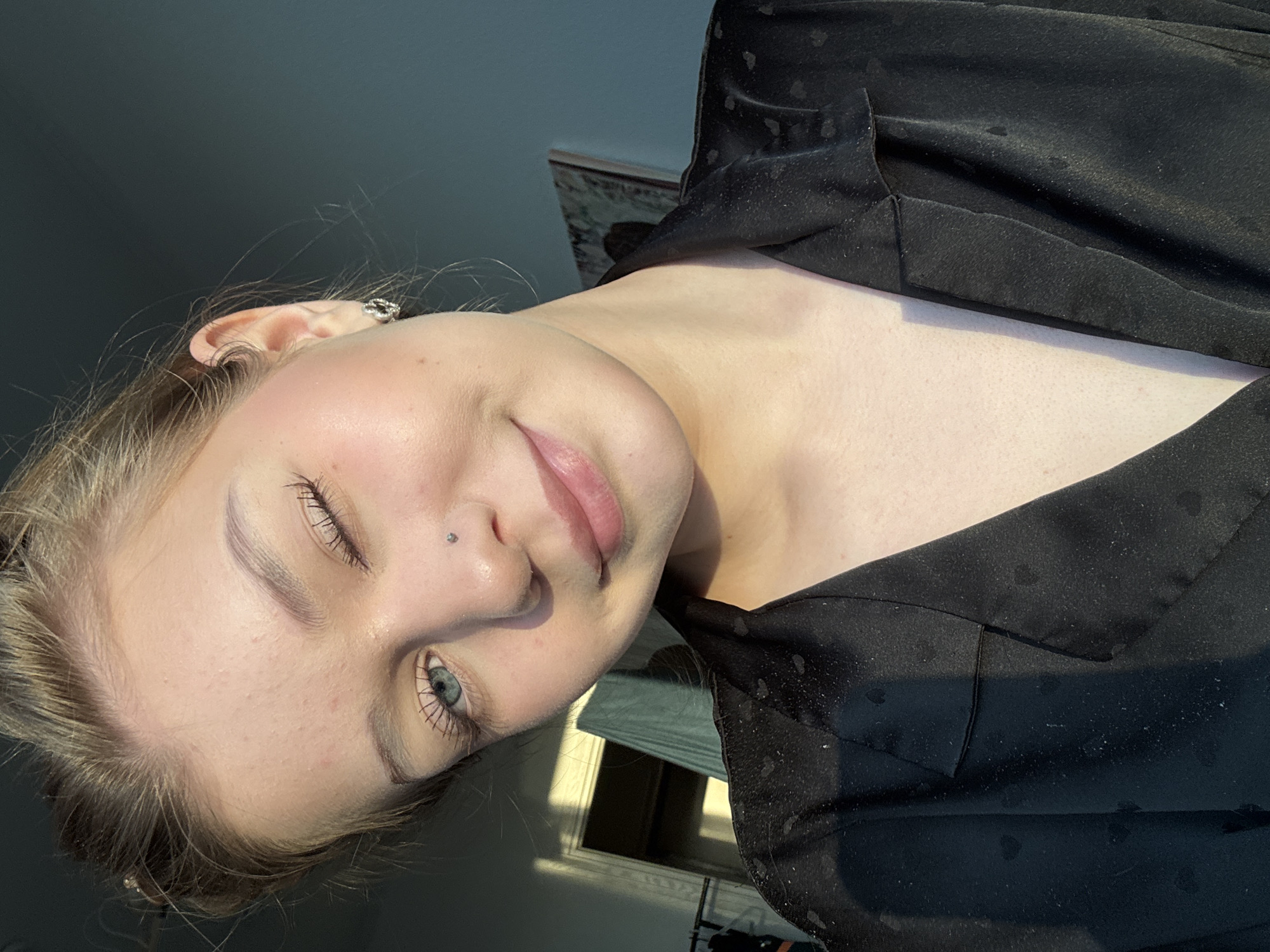 A young woman is depicted in a close-up, angled head and shoulder shot. She has fair skin, light blonde hair pulled back, and light blue eyes. Her lips are a natural pink color. There's a small stud piercing on the left side of her nose. She is wearing a dark-colored garment with a subtle pattern. The background is a dimly lit room. A window with partially open blinds is visible behind her, allowing some sunlight to enter the room. The location of the image is indiscernible as Yekaterinburg, Russia.
