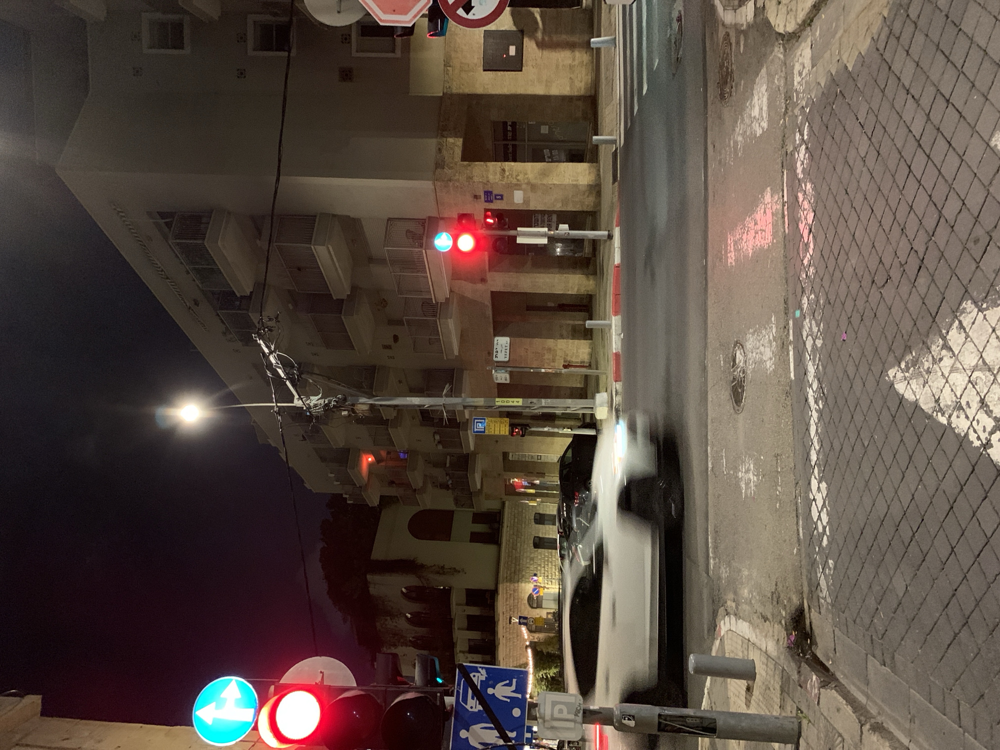 The image shows a street scene at night in Tel Aviv, Israel. A blurred white car is in motion on the right side of the street. Sidewalk tiles with a diamond pattern are also on the right. On the left side of the street, there is a building with balconies. Traffic lights are visible, displaying red and green signals. A street light is present near the middle of the frame. Road signs are also visible on the left.