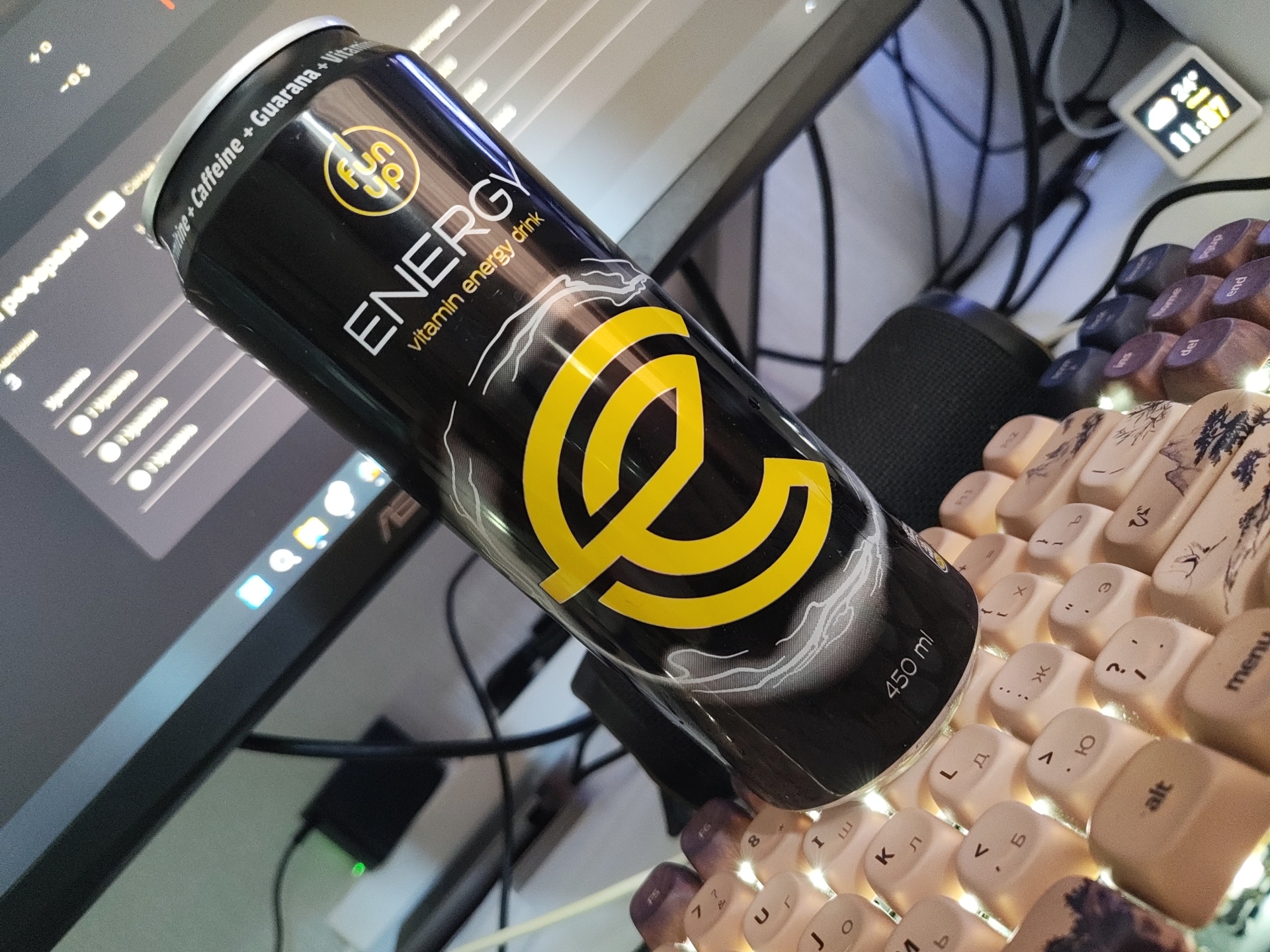 The image shows a close-up of a black energy drink can with a yellow logo and text that reads "ENERGY vitamin energy drink" and "fun up". The can is tilted and rests on a keyboard. The keyboard has illuminated keys with various symbols and Cyrillic letters. In the background, a computer monitor is visible, displaying what appears to be a graphical user interface with icons and text. There is also a small digital device on the right side of the image displaying the time "11:57" and a temperature of "24°". The scene is indoors, likely a desk setup with a computer and peripherals. There are no people or specific landmarks that would indicate the location in Il'inskiy, Russia.
