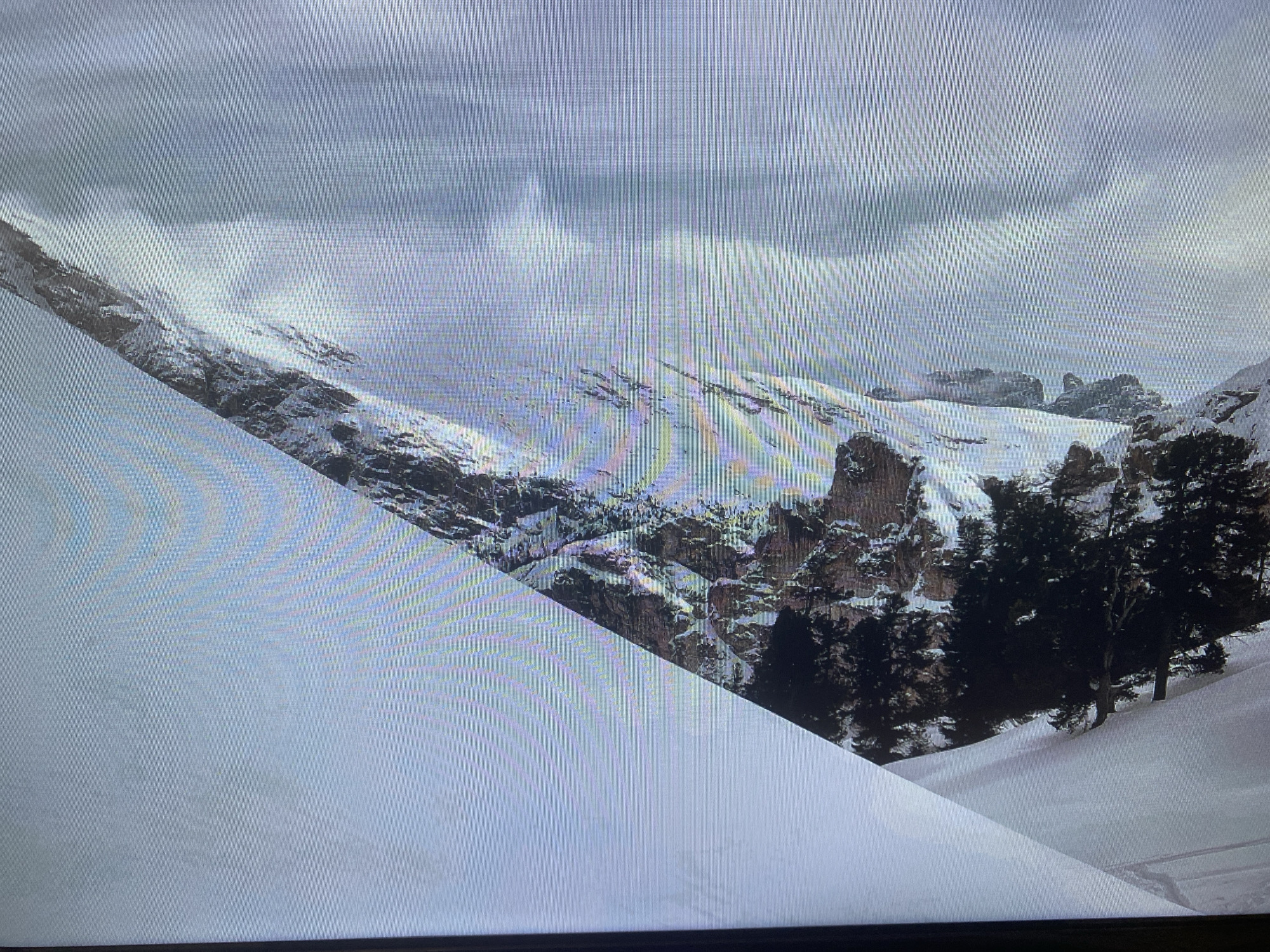 The provided image and its accompanying crops present a expansive, snow-covered mountain landscape under a heavy, overcast sky. The scene is dominated by white snow, grey clouds, dark rock formations, and clusters of evergreen trees.
In the foreground, a large, smooth snow slope occupies the left half of the frame, angling downwards. Towards the bottom right of this slope, subtle tracks, likely from skis or a snowboard, can be faintly observed, indicating prior human activity.
The midground and background reveal rugged, rocky mountains, with their peaks and slopes extensively covered in snow. Dark, coniferous trees are scattered among the rock formations and along the valleys, providing a strong contrast to the white snow. Several distinct, jagged rock outcrops are visible, particularly on the right side of the main image. The sky above is entirely covered by a thick layer of grey clouds, suggesting either an ongoing wintry weather system or just an overcast day.
The time of day appears to be daytime, characterized by a soft, diffused light due to the heavy cloud cover. The weather is clearly cold and wintry, with deep snow blanketing the landscape. There are no visible people or animals, contributing to an atmosphere of remote wilderness and solitude.
A notable technical detail present across all images is a pattern of fine horizontal lines, indicating that these are photographs taken of a digital screen rather than direct captures of the scene. No discernible text is visible within the image.
The visual evidence of high, rocky, snow-capped mountains is inconsistent with the provided location context of "Saint Petersburg, Russia," which is not known for such mountainous terrain.