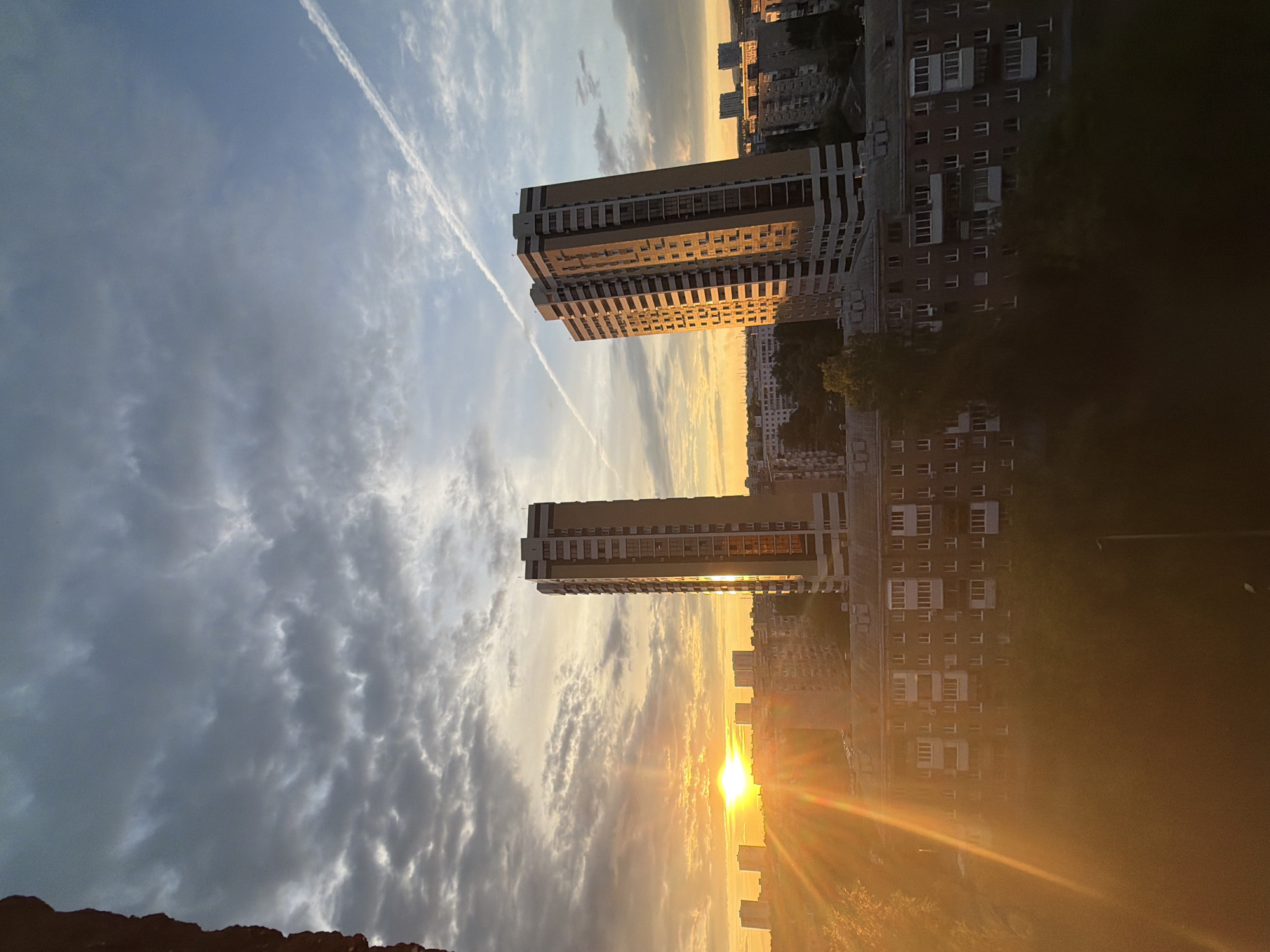 The image, correctly oriented, captures an urban landscape at either sunrise or sunset, characterized by dramatic lighting and architectural structures. A brilliant golden sun is low on the right horizon, casting intense rays that create a strong lens flare and bathe the scene in warm, golden light.
Two prominent, modern high-rise apartment buildings stand tall in the mid-ground, their upper floors reflecting the sun's glow. Surrounding them and extending into the distance is a dense cityscape comprised of numerous other apartment blocks, some partially obscured by shadows. The foreground on the bottom-left shows a dark, indistinct rocky or elevated surface.
The sky is a striking mix of deep blues, greys, and luminous oranges and yellows, showcasing various cloud formations from wispy streaks to textured clusters. A distinct, white contrail from an aircraft cuts diagonally across the sky from the top-left, passing between the two main towers. Patches of dark green vegetation, likely trees or bushes, are visible as silhouettes in the shadowed areas near some buildings. The scene conveys a serene yet grand atmosphere, highlighting the contrast between urban architecture and the natural beauty of the golden hour. No human activity or visible text is present.
Location context: Unknown, Unknown