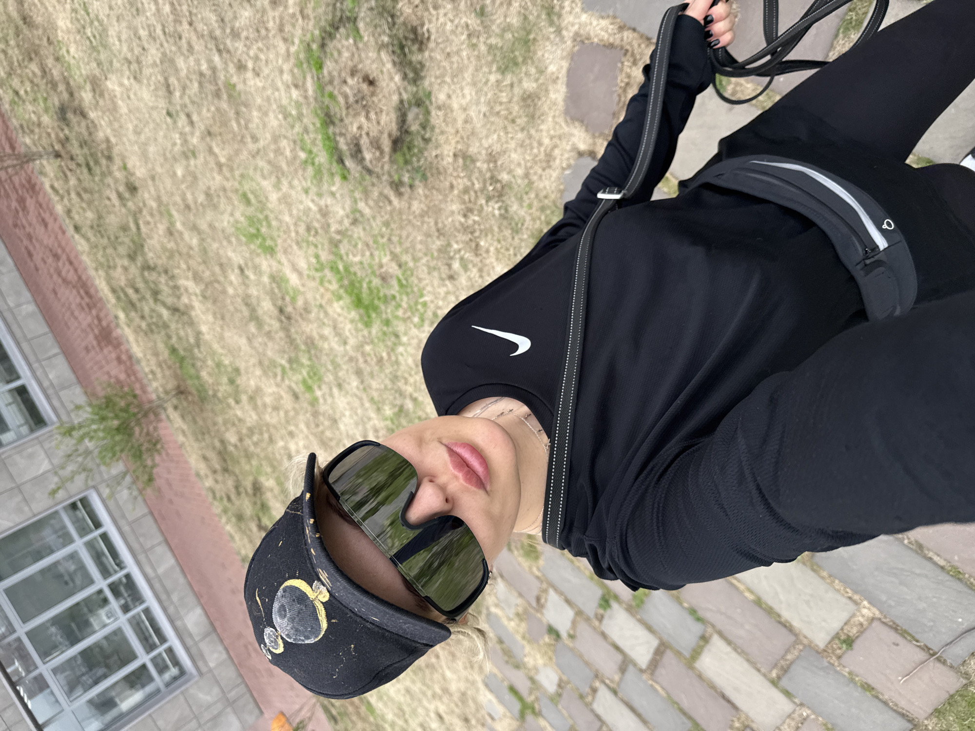A person wearing black athletic wear, a cap, and sunglasses stands on a stone-paved area near a lawn and a building with windows. The location is identified as Krasnoyarsk, Russia. The person holds a dog leash.