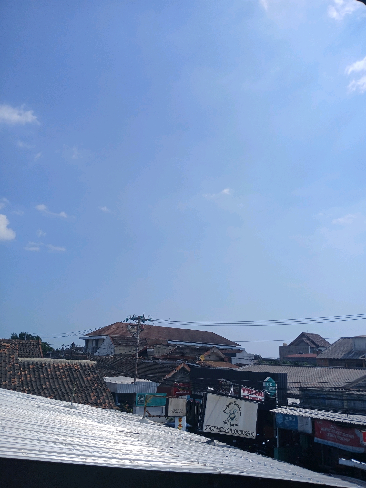This image captures a view of a densely populated urban area in Depok, Indonesia, under a clear blue sky with scattered white clouds. The foreground is dominated by a corrugated metal roof, reflecting the sunlight. Behind this, a jumble of buildings with various roofing materials, including clay tiles and darker metal sheets, are visible.
Several signs are present on the buildings, indicating commercial activity. One prominent sign, written in Indonesian, reads "Penyetan Ibu Suradi," likely advertising a local eatery. Other signs are partially visible and appear to advertise services such as "Salon" and possibly shipping or delivery services, indicated by "J&T" and "KIRIM PAKET." A utility pole with electrical wires runs across the middle ground, a common sight in urban Indonesian landscapes. The overall impression is of a bustling, everyday street scene in an Indonesian city, likely a commercial district or a mixed residential-commercial area. There are no people clearly visible in the image.