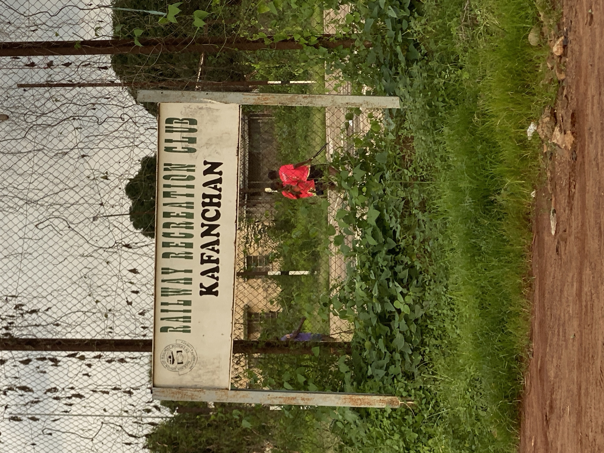 The image depicts the entrance to the Railway Recreation Club in Kafanchan, Maigizo, Nigeria. A white sign with green lettering reads "RAILWAY RECREATION CLUB" above "KAFANCHAN." To the left of the text, there is a circular emblem with a train and the words "UNION OF RAILWAYMEN NIGERIA." The sign is supported by two metal posts, one on the left and one on the right. Behind the sign, a chain-link fence runs horizontally across the frame.
Within the fenced area, two individuals wearing bright red shirts and dark shorts are walking. One is carrying what appears to be a long, thin object, possibly a stick or a piece of sporting equipment. Another person in a purple garment is partially visible further to the left, behind a fence post. In the background, a beige building with windows is visible, surrounded by lush green vegetation. Overcast, cloudy skies are visible above the fence and trees. The foreground shows a dirt path and more overgrown greenery.