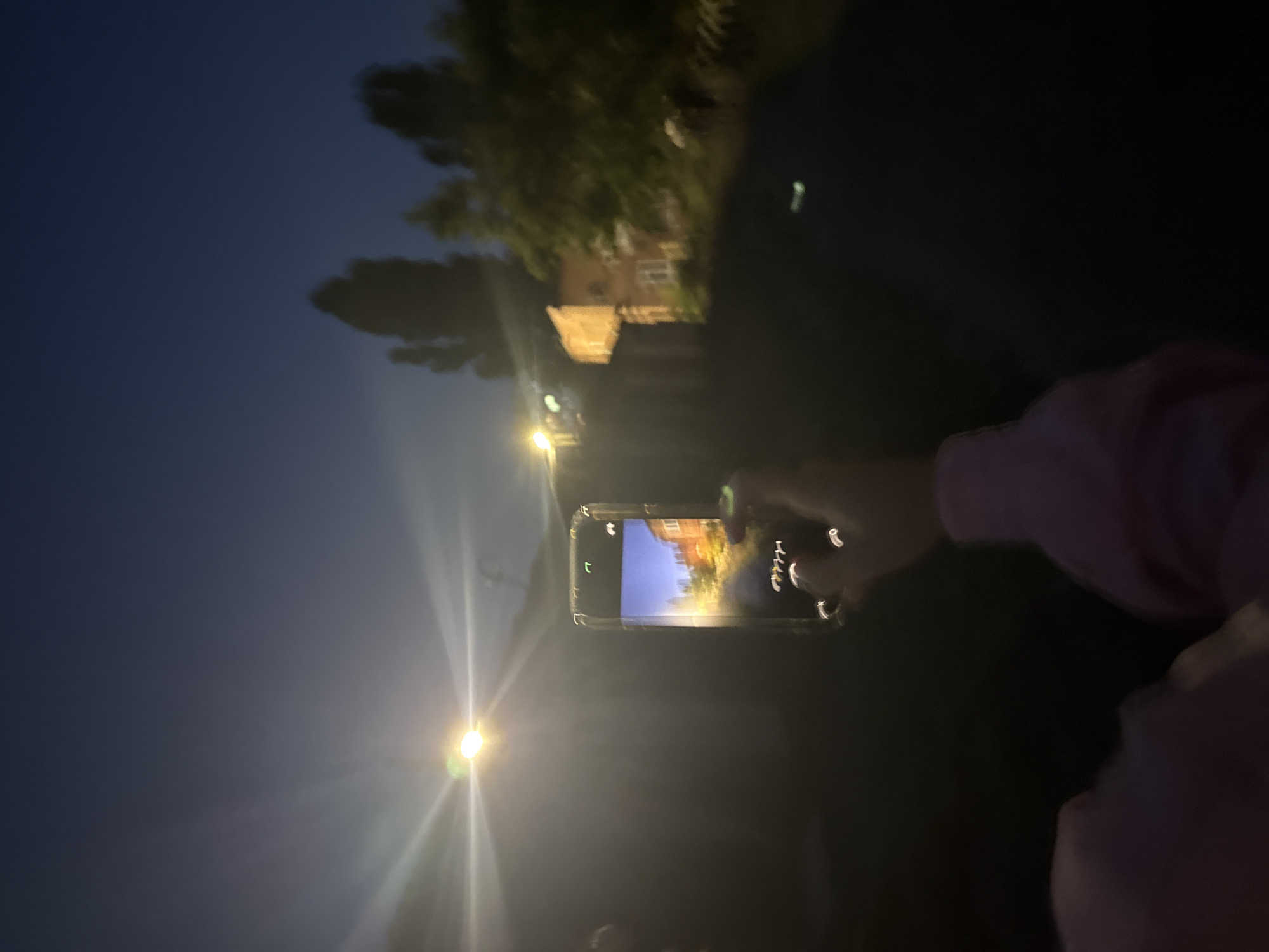 An out-of-focus, vertical, medium shot captures a hand holding a smartphone in the foreground, with the phone's screen displaying a brightly lit building. The background is a dark outdoor scene with a streetlamp emitting a strong glare. In the distance, a few buildings and trees are visible against a dark blue twilight sky. The overall impression is of a photograph being taken at dusk or night, possibly in a residential area.