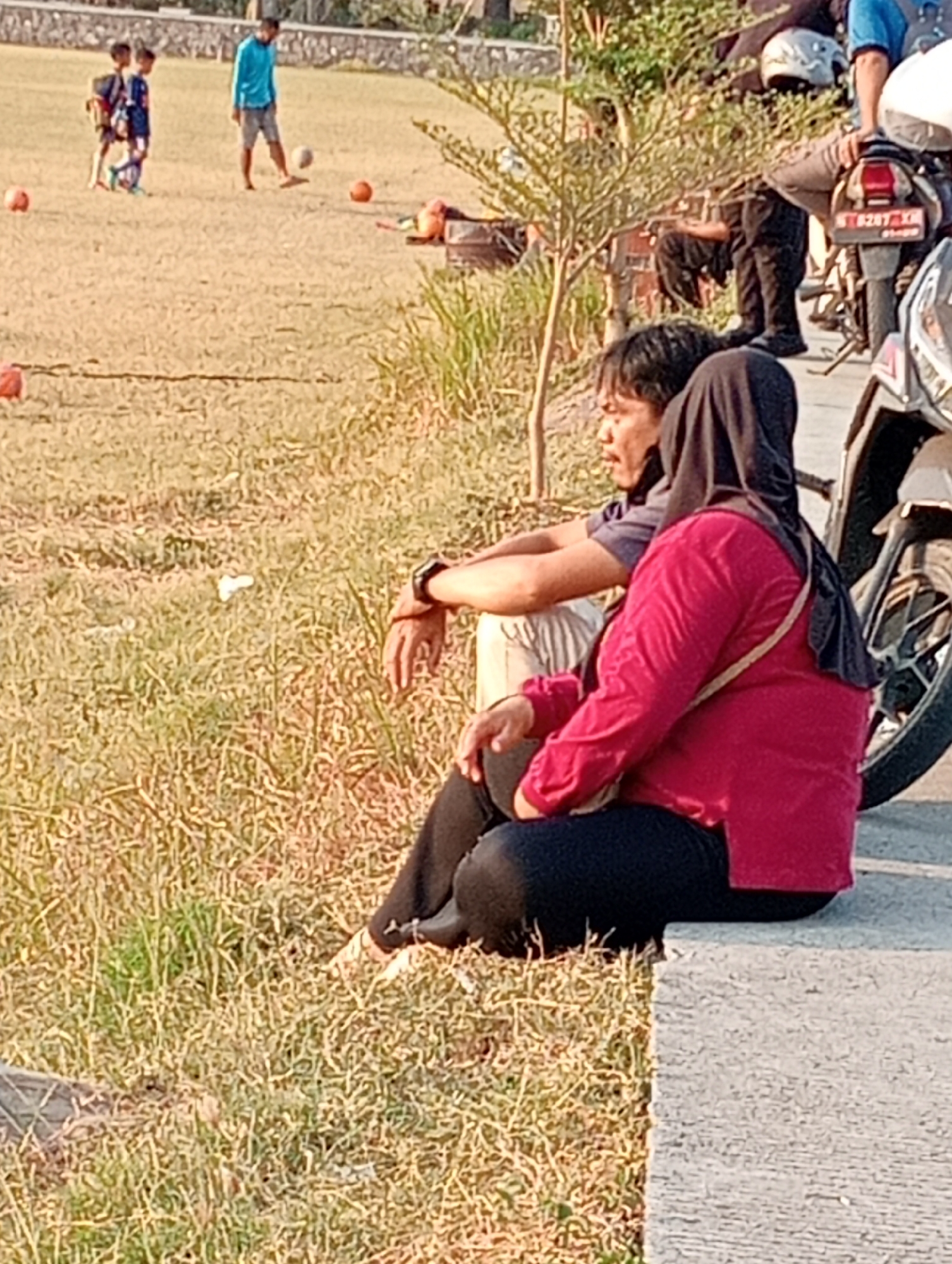 In Kenep, Indonesia, two people sit on a concrete ledge overlooking a grassy field where three children are playing soccer. The male, on the left, wears a purple shirt and light pants. The female, on the right, is dressed in a red long-sleeved shirt and black pants, with a dark hijab. A motorcycle is parked to their right, partially obscuring a brick wall in the background. The scene is set during the day, with warm sunlight illuminating the dry grass and the figures.