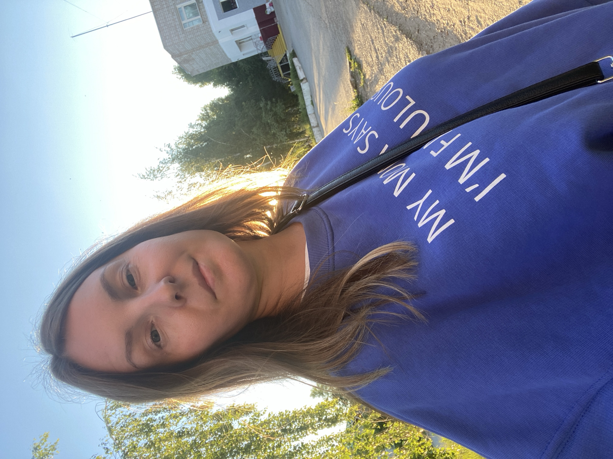 A woman with light brown hair is wearing a blue sweatshirt with white lettering, standing outdoors. The text on her sweatshirt reads "MY MIND SAYS I'M F*CKING FABULOUS." She has a black purse strap across her chest. In the background, there's a building with white and gray exterior elements and green trees. The location is Noviy Urengoy, Russia. The photo is taken during daylight hours with sunlight present.
