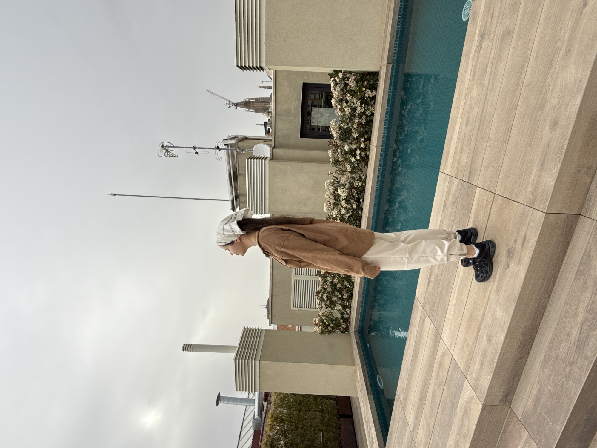 The image captures a person standing beside a pool on a rooftop in Barcelona, Spain. The person is wearing a brown jacket, light-colored pants, a patterned cap, and black sneakers. They are looking down towards the pool. The pool is lined with light-colored tiles, and the surrounding area is paved with wooden planks.
In the background, a cityscape is visible, including buildings, a construction crane, and antennas. The sky is overcast. A row of flowers are also visible.