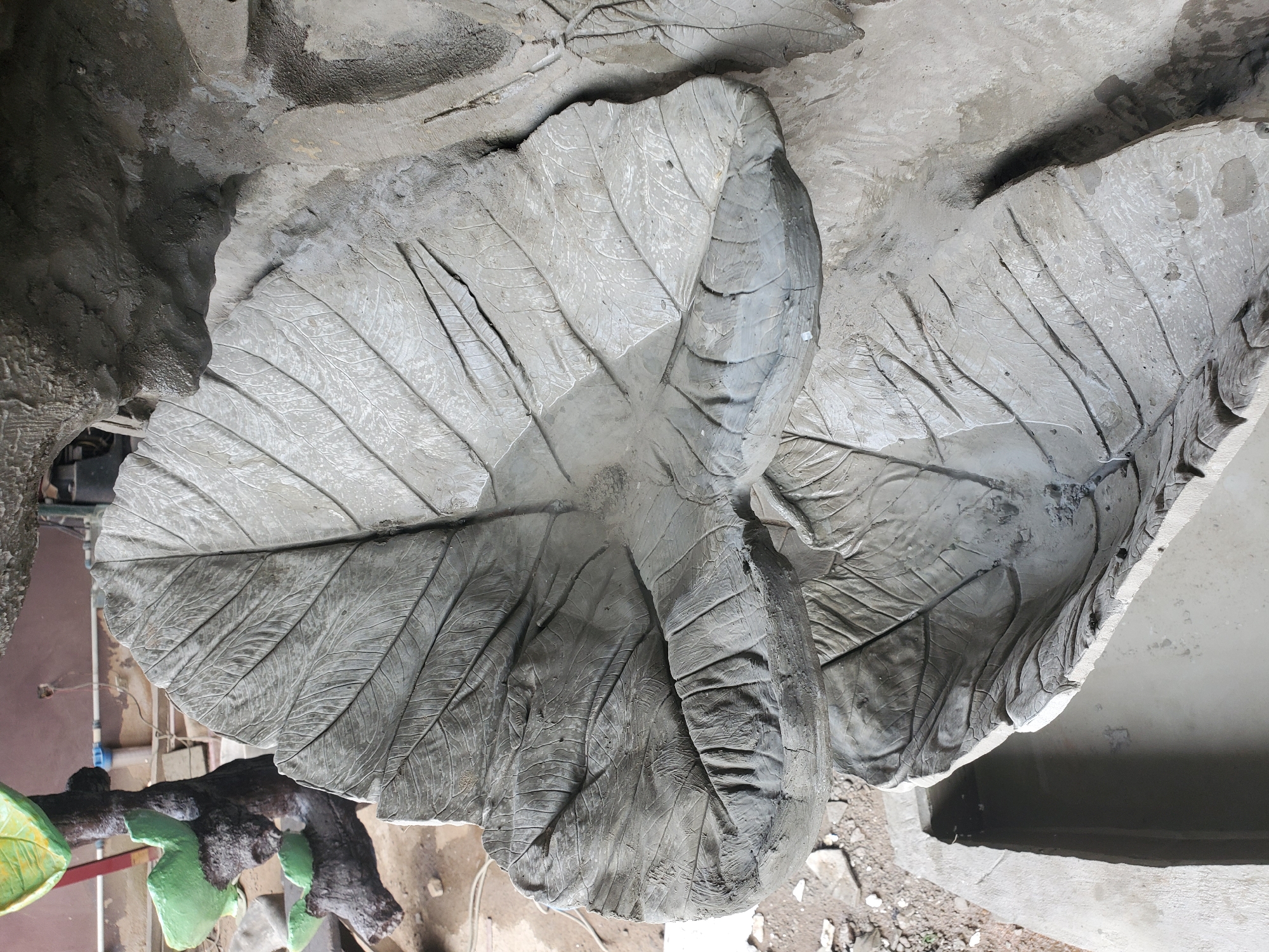 The image displays a close-up, overhead view of a decorative concrete structure resembling large, textured leaves. The primary subject is a large, grey leaf formation, with a smaller, similar leaf structure below it. The leaves exhibit prominent vein patterns and a rough, unfinished texture, indicative of concrete casting. The upper leaf appears to be a basin, holding a small amount of murky water. The surrounding environment is rough and appears to be an outdoor or workshop setting, with loose gravel and building materials visible in the background, including what looks like plumbing fixtures and a dark, cylindrical object. The overall impression is that this is a work in progress for a garden feature or fountain. The provided context of Jos, Nigeria, suggests this scene is located within that city.