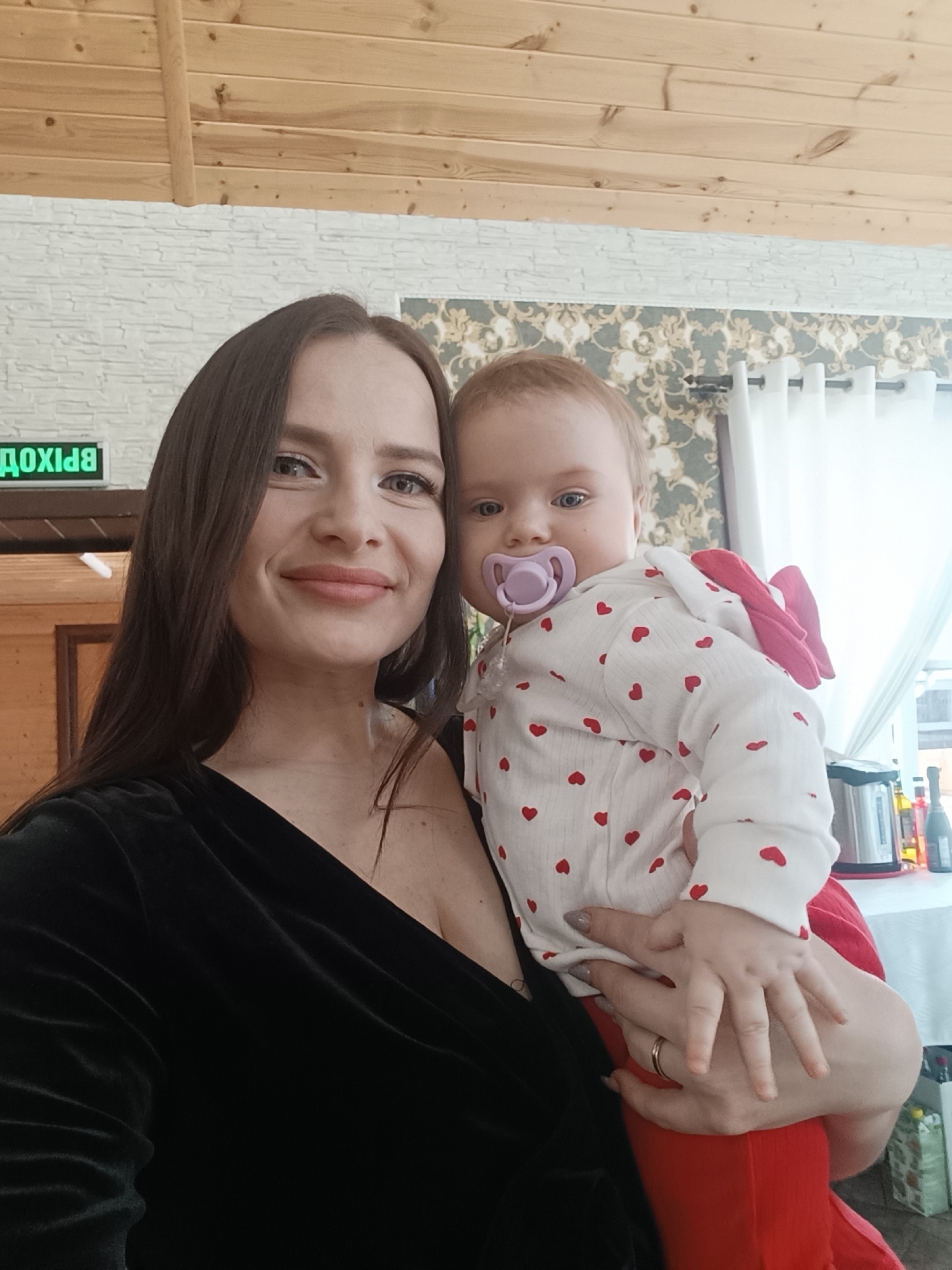 A woman is holding a baby in front of a background composed of wood paneling, a decorative wall, and a window. The woman has long, dark brown hair, light skin, and is wearing a black velvet dress with a low neckline. The baby is wearing a white long-sleeved shirt patterned with red hearts and red pants and has a purple pacifier in its mouth. The woman's left hand is holding the baby. Other objects visible in the background include an exit sign (in Russian), a kettle, and bottles on a table by the window. The location is in the city of Chaikovsky, Russia.