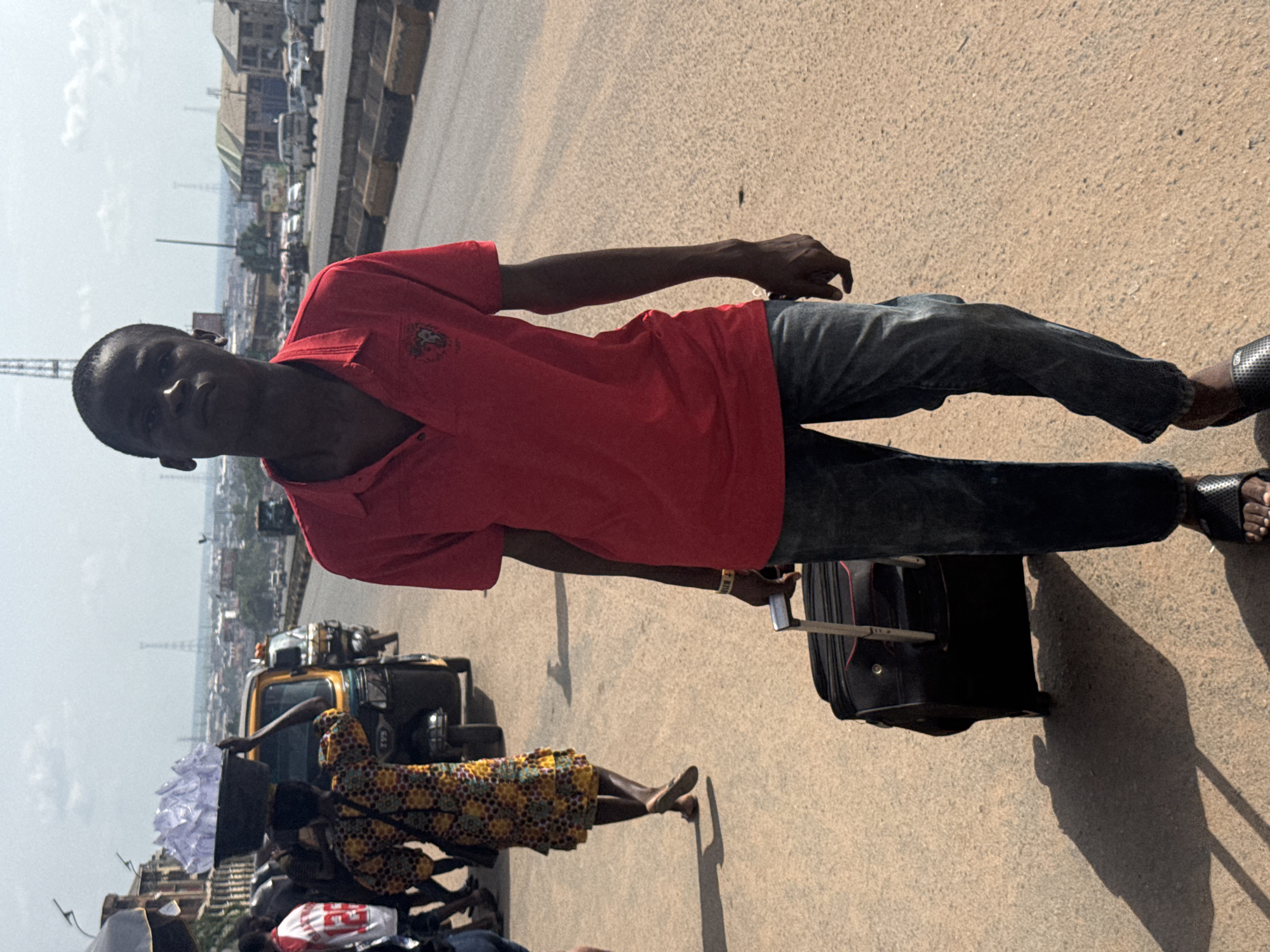 A man in a red short-sleeved shirt, jeans, and sandals pulls a black suitcase on a paved surface. He holds a phone in his left hand. In the background, a yellow tractor-like vehicle and people are visible, along with other vehicles in the distance. The location appears to be a bridge or elevated road in Onitsha, Nigeria, based on the urban environment.