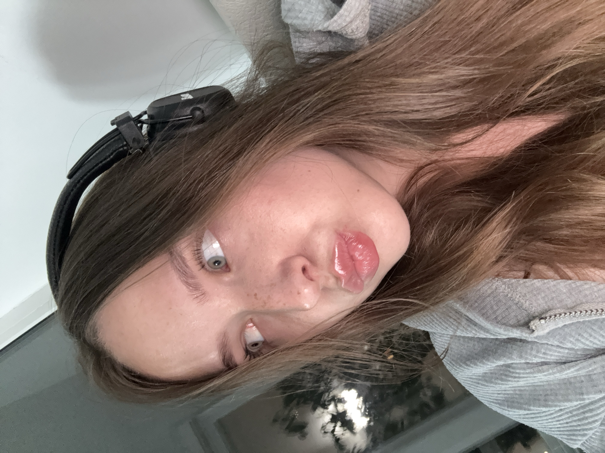 This is a close-up, selfie-style portrait of a young woman wearing black headphones. She has long, light brown hair, fair skin with freckles, and is looking to her left with her lips pursed. She is wearing a grey ribbed hooded jacket. The background includes a window with reflections of trees and lights, suggesting an indoor setting, possibly an apartment. There is no specific indication of location in the city of Chaikovsky, Russia within the image itself.