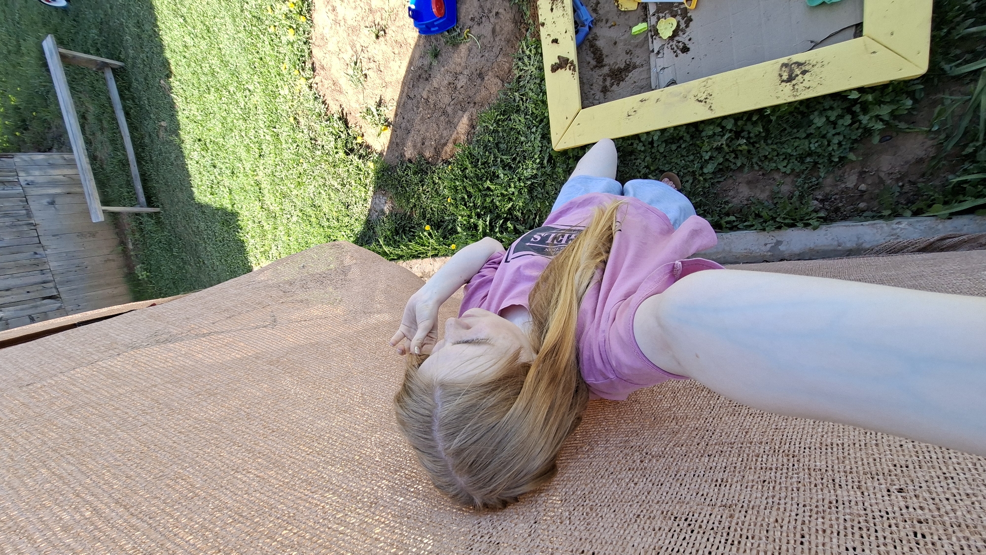 A person with long blonde hair is lying on a tan woven surface outdoors. The person is wearing a pink t-shirt and light-colored shorts. To the left of the person is a wooden structure and grass, and to the right, there's a sandbox with yellow frame and grassy area surrounding it. The location is outdoors and seems like a backyard. No information suggests the location is in the city of Большой Букор, Russia.