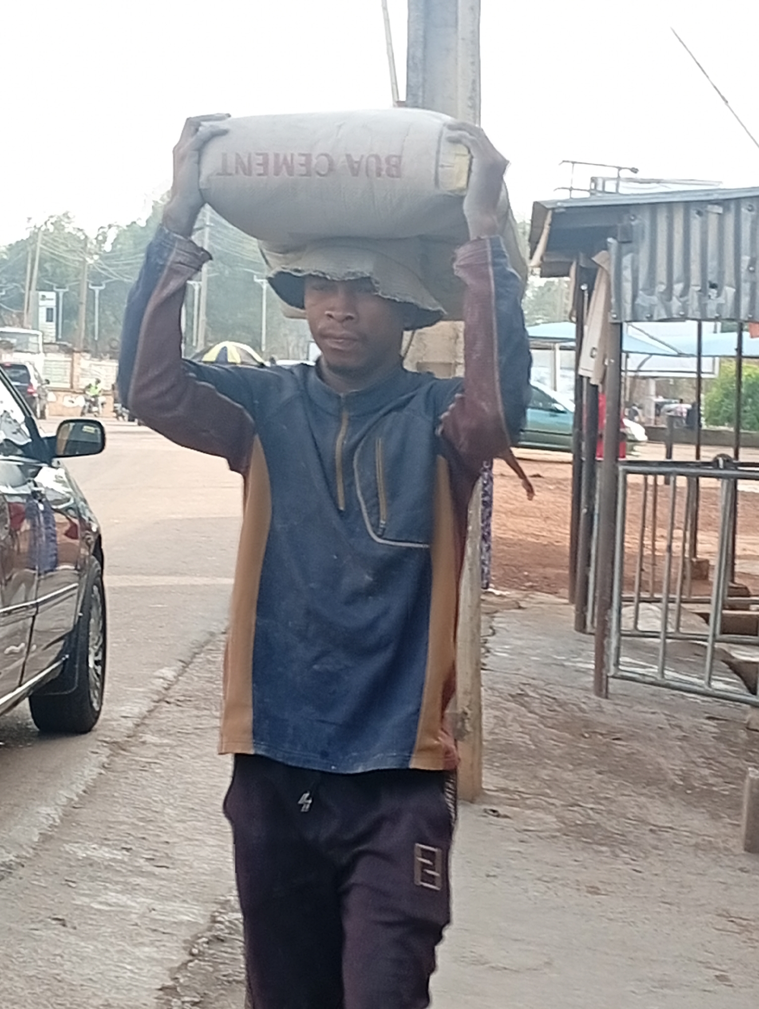 A man carries a bag of "BUA CEMENT" on his head in Gura Top, Nigeria. He is wearing a long-sleeved, two-toned shirt and dark pants. He is standing on a street next to a car. In the background, there are buildings and other vehicles.