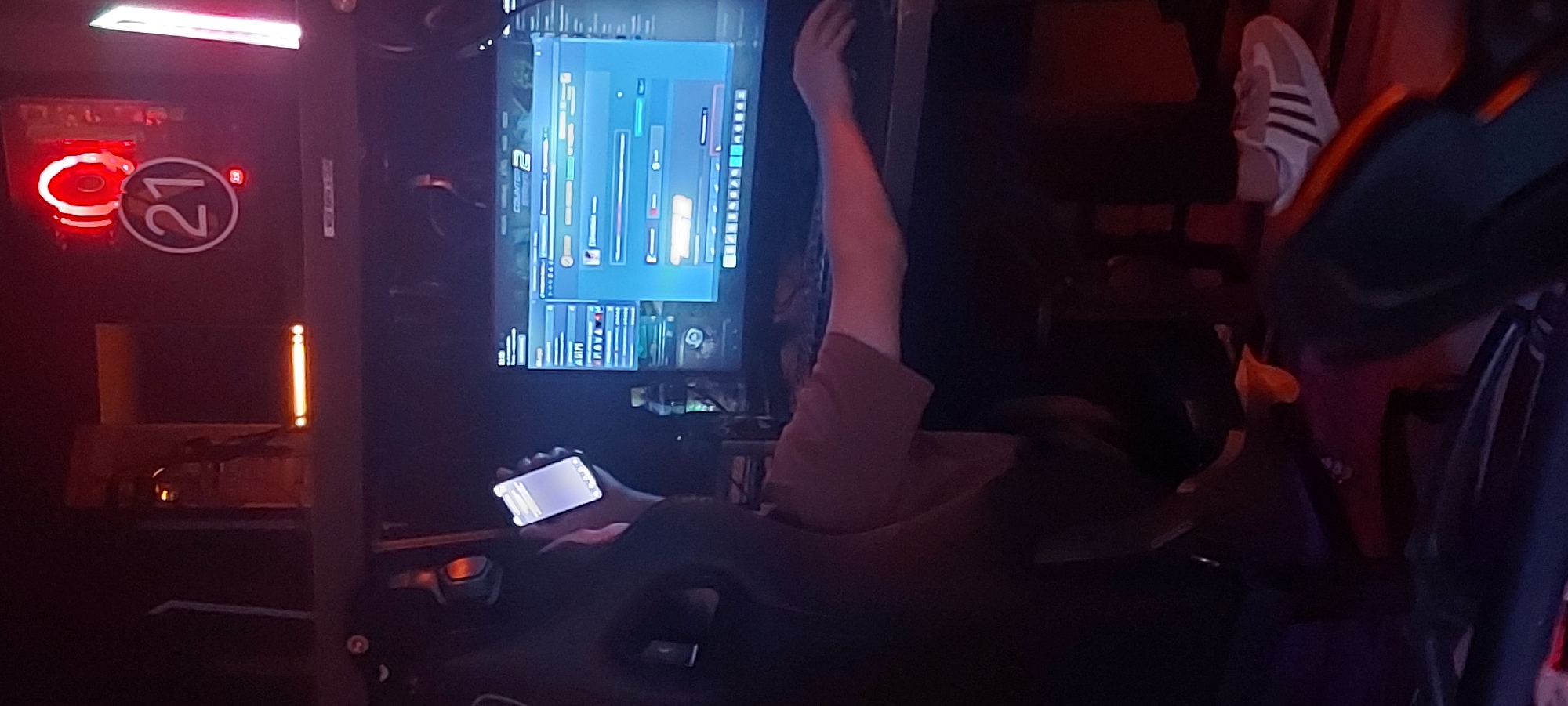 The image shows an interior scene. A person is sitting in a racing seat with their arm raised near a computer screen displaying a graphic interface that appears to be part of a simulation or gaming setup. The person is holding a phone in their other hand. There's a red circular light with the number "21" next to it. A pair of Adidas shoes are visible on the upper right side of the frame. There is a yellow light on the left side of the frame. The scene is generally dark with various elements of a gaming or simulation setup visible. There is no explicit indication of the city of Tashkent, Uzbekistan.