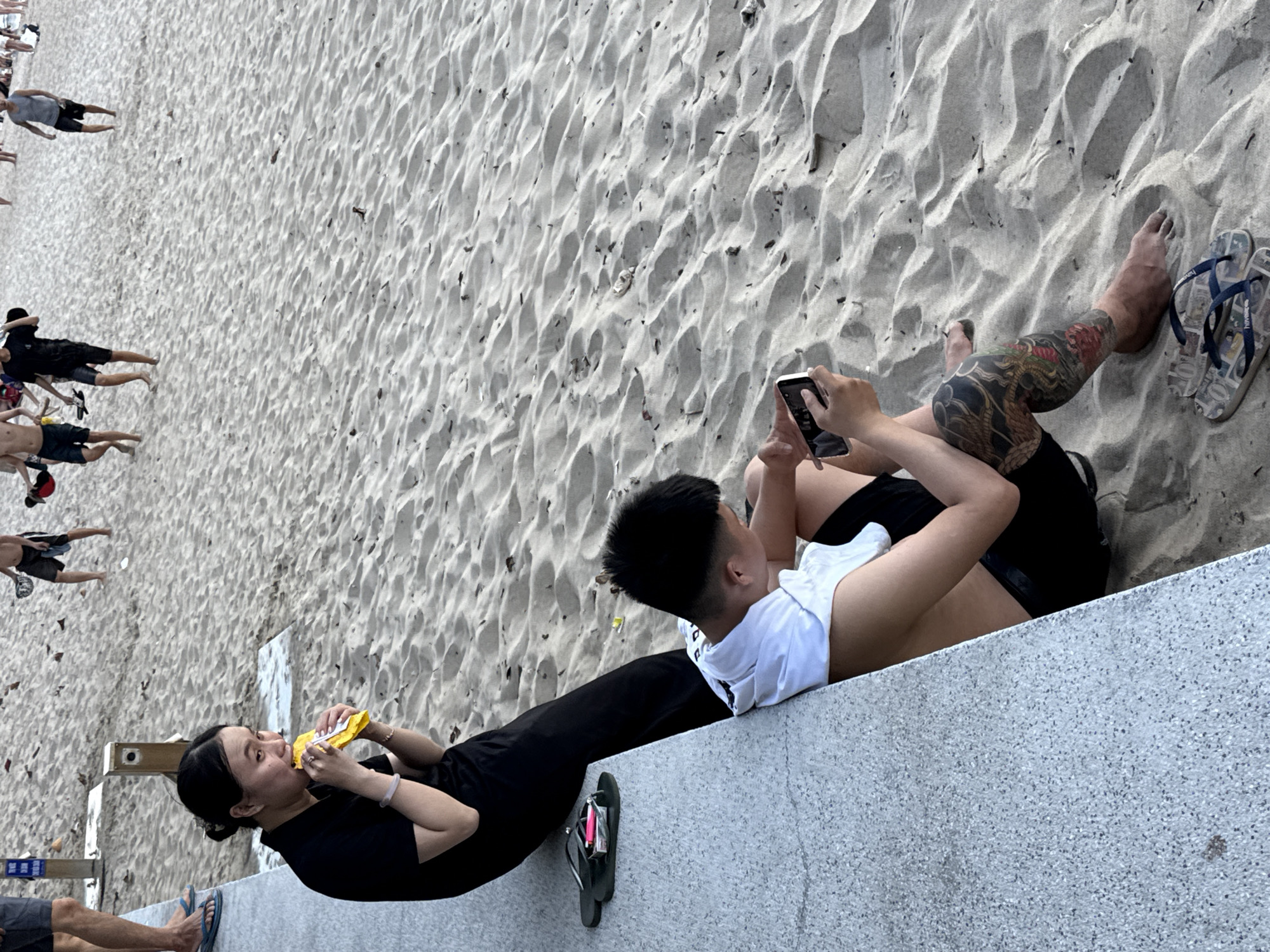 The image depicts a beach scene in Nha Trang Ward, Vietnam. In the foreground, a woman in a black top and pants is seated, eating from a yellow packet. Beside her, a man in a white t-shirt and black shorts is sitting, looking at his phone. He has a large tattoo on his leg. Flip-flops are visible on the sand near the man and also by the woman. In the background, other people are walking on the beach. The environment is sandy with ripples indicating the tide or wind.