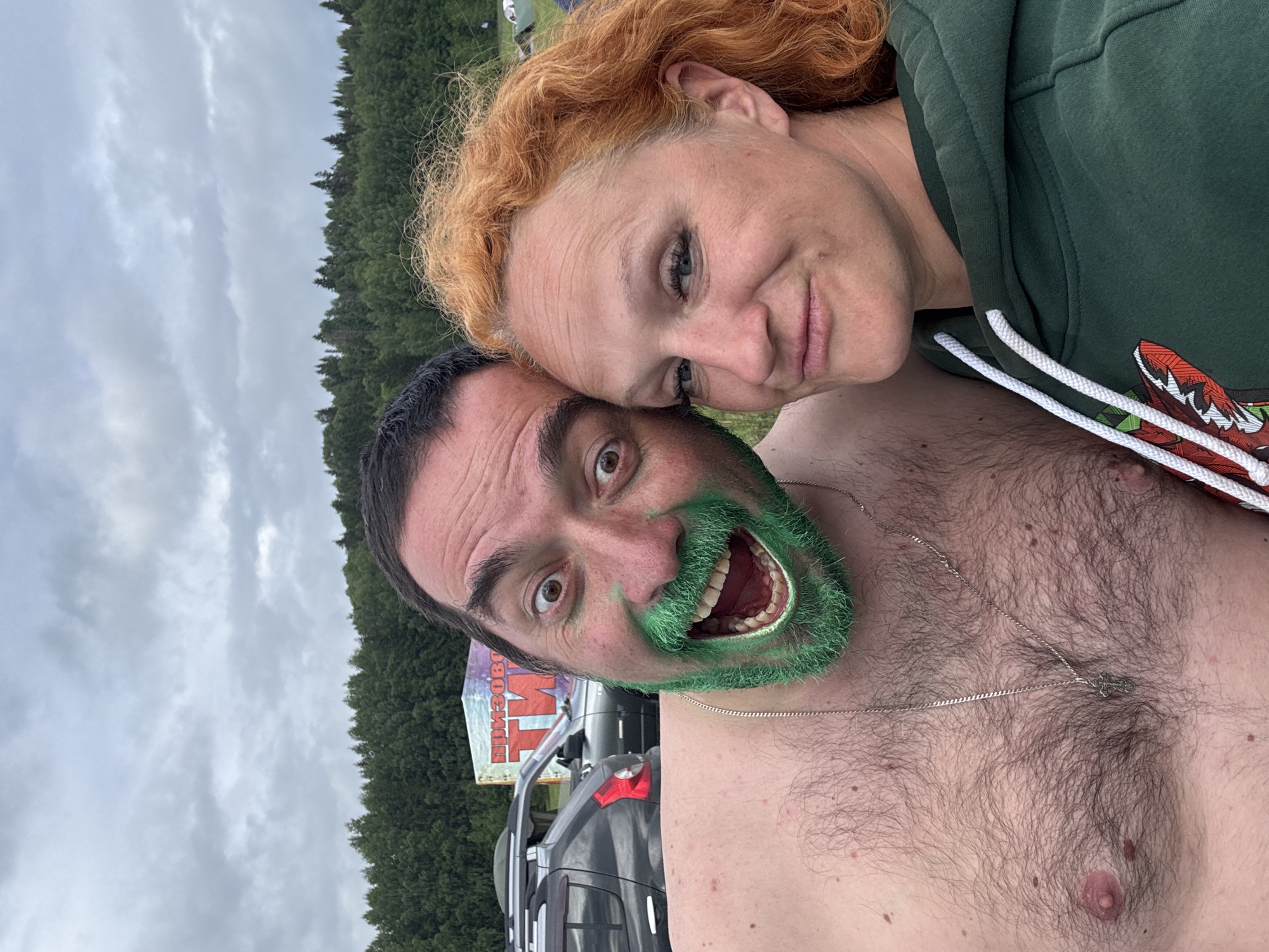 This close-up photograph features two individuals, a man and a woman, captured in a playful outdoor setting. The woman, positioned slightly above and behind the man, has wavy reddish-orange hair and wears a dark green hooded sweatshirt. She leans her head against the man's, offering a soft smile with light-colored eyes looking towards the viewer.
The man, in the foreground, has dark hair and a full beard painted a vibrant green, with matching green paint around his face. He displays an exaggerated, joyful expression with a wide-open mouth revealing his teeth and tongue, and widely open eyes. He is shirtless, showing a hairy chest adorned with a silver chain featuring a cross pendant.
The background reveals a cloudy sky stretching above a dense line of dark green evergreen trees, suggesting a forested or natural environment. Partially visible behind the man and below the tree line are several parked dark-colored vehicles, possibly SUVs or trucks. A large sign with Cyrillic text, reading "ПРИВОЛЖЬЕ ТМ" (Povolzhye TM), is discernible behind the vehicles. The overcast sky indicates it is daytime. The green face paint and joyful expressions suggest a festive or celebratory outdoor event, likely taking place in Большая Кивара, Russia, as per the location context.