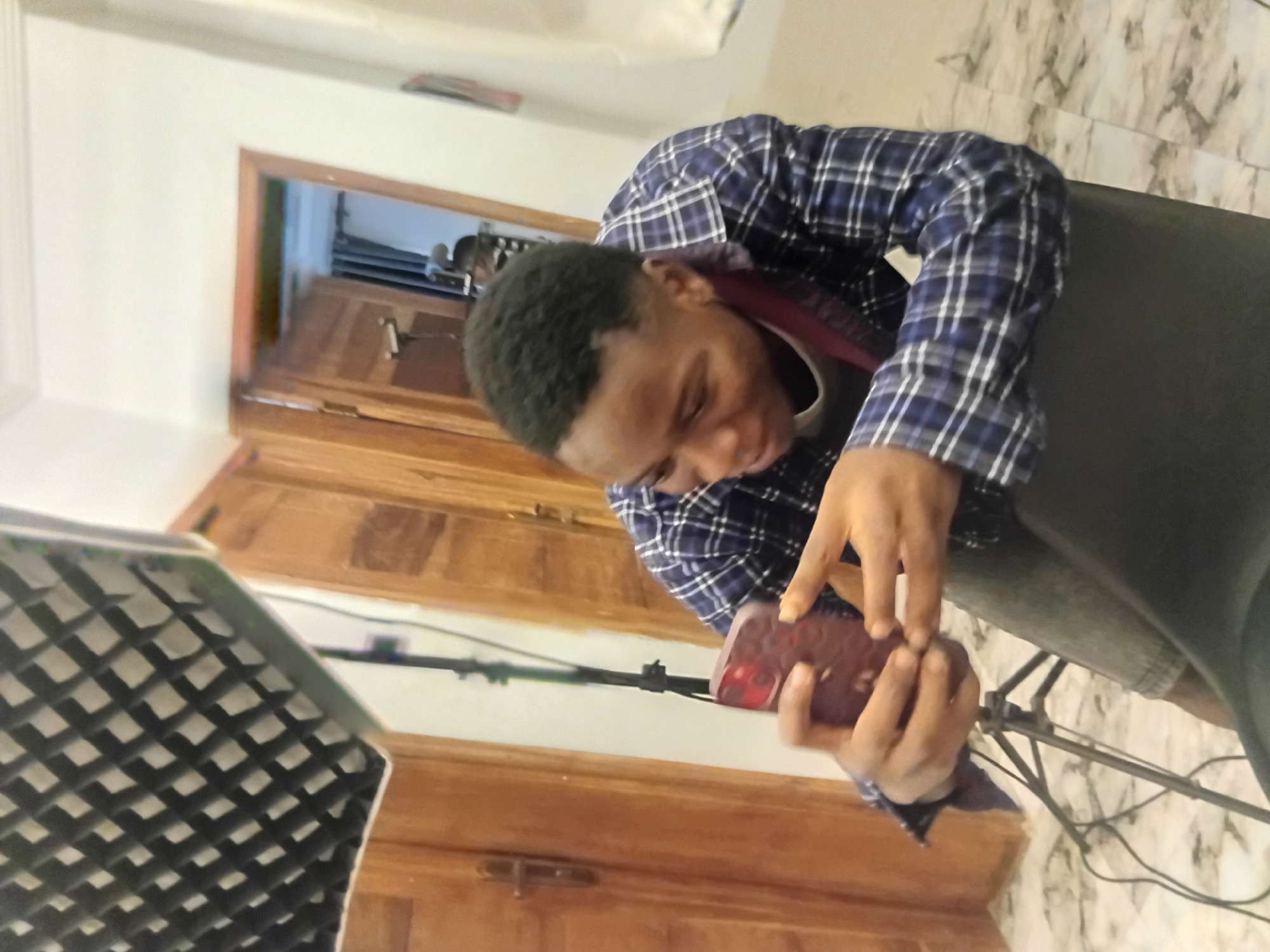 A young man in a blue and white checkered shirt sits in a black chair, holding a red smartphone. He appears to be using the phone with both hands, his focus directed downwards. The scene is illuminated by a softbox light with a honeycomb grid, positioned to the left. In the background, a set of wooden doors leads to another room. The environment includes patterned floor tiles and white walls. While the location is specified as Ikorodu, Nigeria, this is not visibly verifiable from the image.
