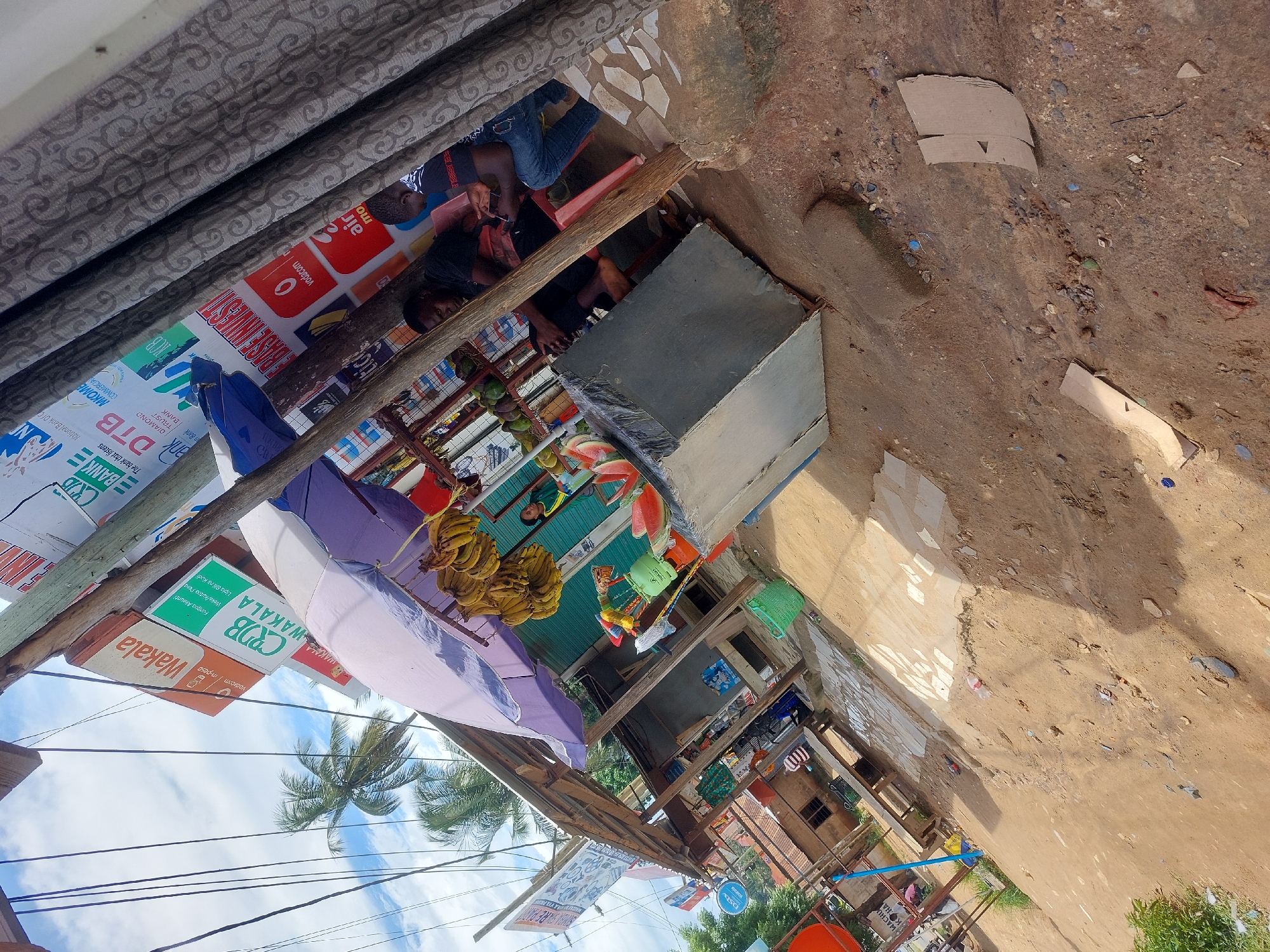 The image captures a street scene in Dar es-Salaam, Tanzania. The scene is dominated by a collection of small businesses operating from open-fronted stalls or shops, evidenced by multiple signage, including that of vodacom Im-pesa, CRDB Wakala, and DTB Bank. These businesses appear to be offering banking or mobile money services. Bunches of bananas hang prominently, indicating a food vendor or market presence. Several people are visible, including individuals sitting or standing near the stalls. Utility poles and power lines run across the sky, partially obscured by palm trees and cloud cover. The street surface is dirt or sand-covered, and the building materials suggest a mix of permanent and temporary constructions. A purple umbrella is used as a sun shade.