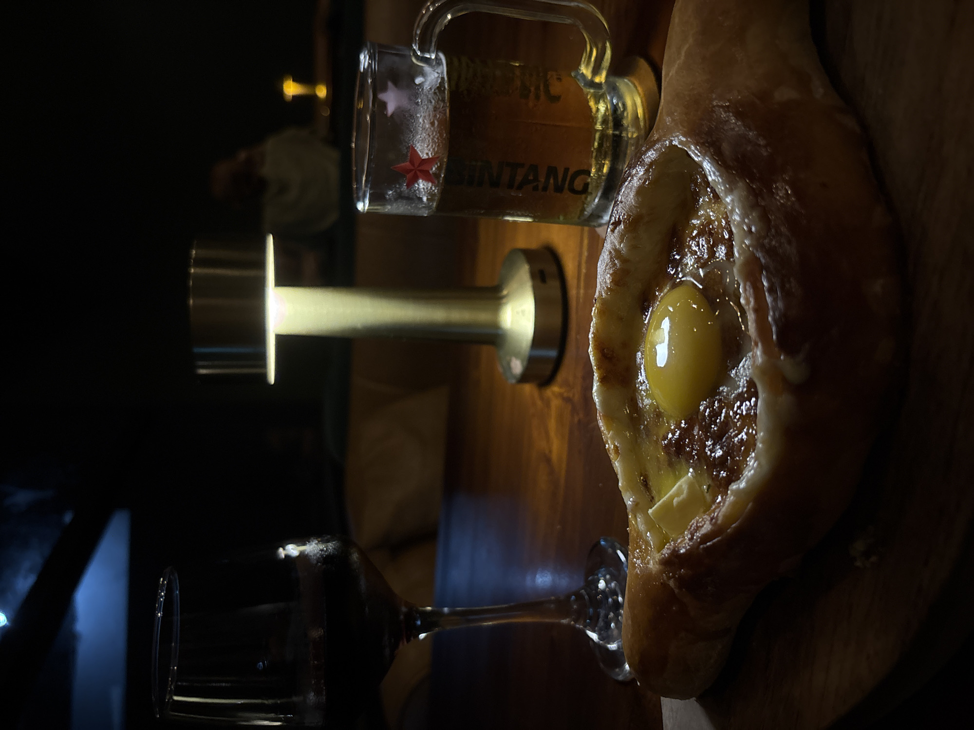 The image depicts a khachapuri, a beer mug labeled "Bintang," and a wine glass on a dark wooden surface. The khachapuri contains a visible egg yolk. The beer mug is partially filled with liquid. A lamp with a cylindrical shade is present. The location is likely Kuta Selatan, Indonesia, given the Bintang beer branding.