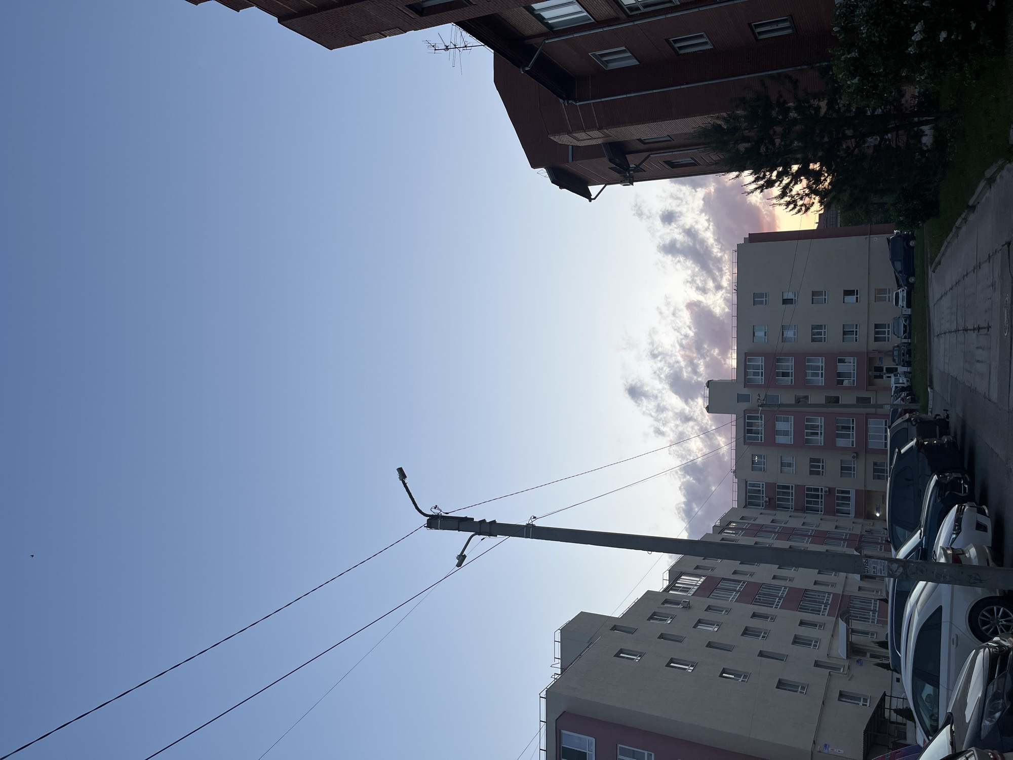 The image shows a street scene in Chaikovsky, Russia. The view is angled upwards, showing a blue sky with some clouds. Two buildings flank the top edges of the frame; a red brick building on the upper left, and a lighter colored apartment building on the upper right with some trees. A tall, gray concrete utility pole stretches from the bottom left to the center of the frame, carrying overhead power lines. A line of parked cars is visible on the right side of the image, parallel to the building, suggesting a street or parking area. The angle of the shot, taken from below, and the presence of buildings on both sides suggest an alleyway or narrow street.
