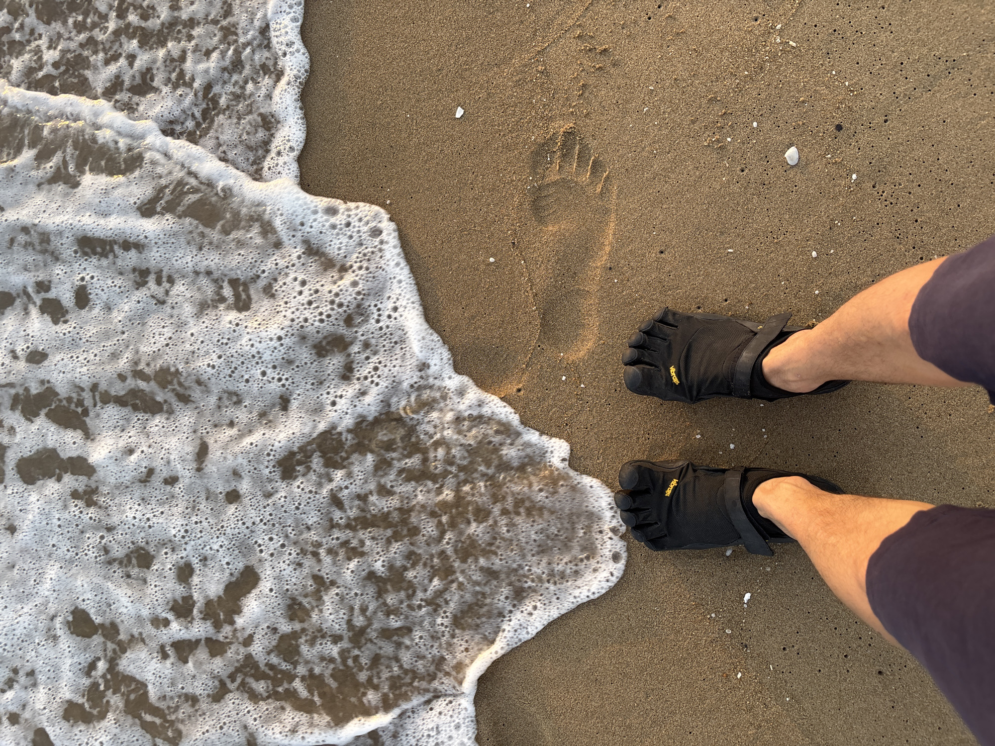 The image captures a low-angle, overhead view of a person's feet wearing black Vibram FiveFingers shoes on a sandy beach. The shoes have individual toe compartments and adjustable straps. The person is wearing dark blue shorts. The sand is a light brown color with small shells scattered across it. A footprint is visible in the sand to the right of the person's feet. The edge of a gentle wave with white foam is encroaching on the sand from the top left of the frame, indicating a beach in Pattaya City, Thailand. The lighting suggests it is either early morning or late afternoon.