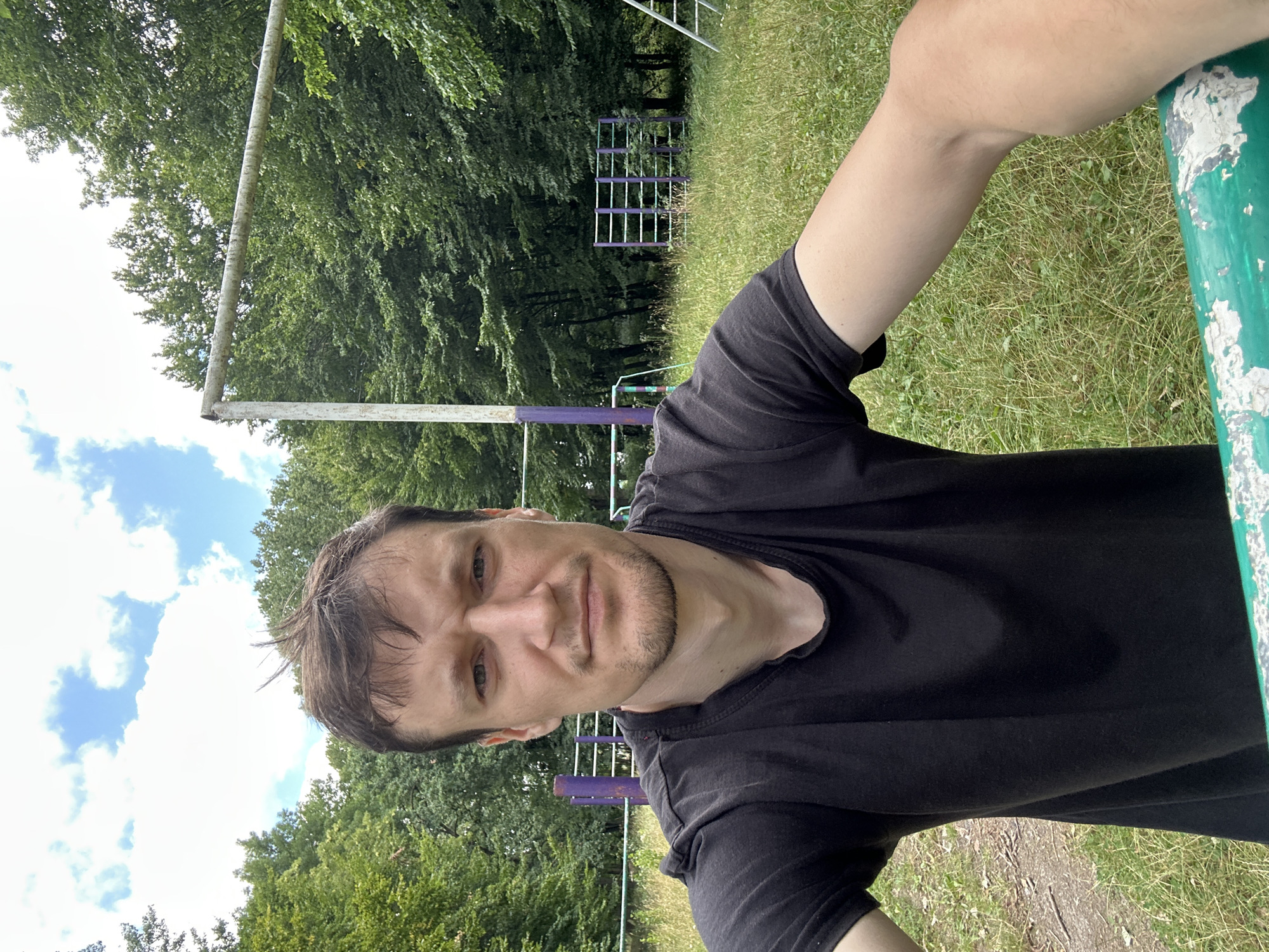 A young man with brown hair and light skin is taking a selfie outdoors. He is wearing a black t-shirt. He is standing in front of a grassy area with trees in the background. There are also some outdoor exercise equipment visible, including bars and a horizontal bar structure. The sky is blue with white clouds. The overall impression is that the man is at an outdoor gym or park. The image quality is good, with clear details and natural lighting. The location is identified as Makiv, Ukraine based on the provided metadata.