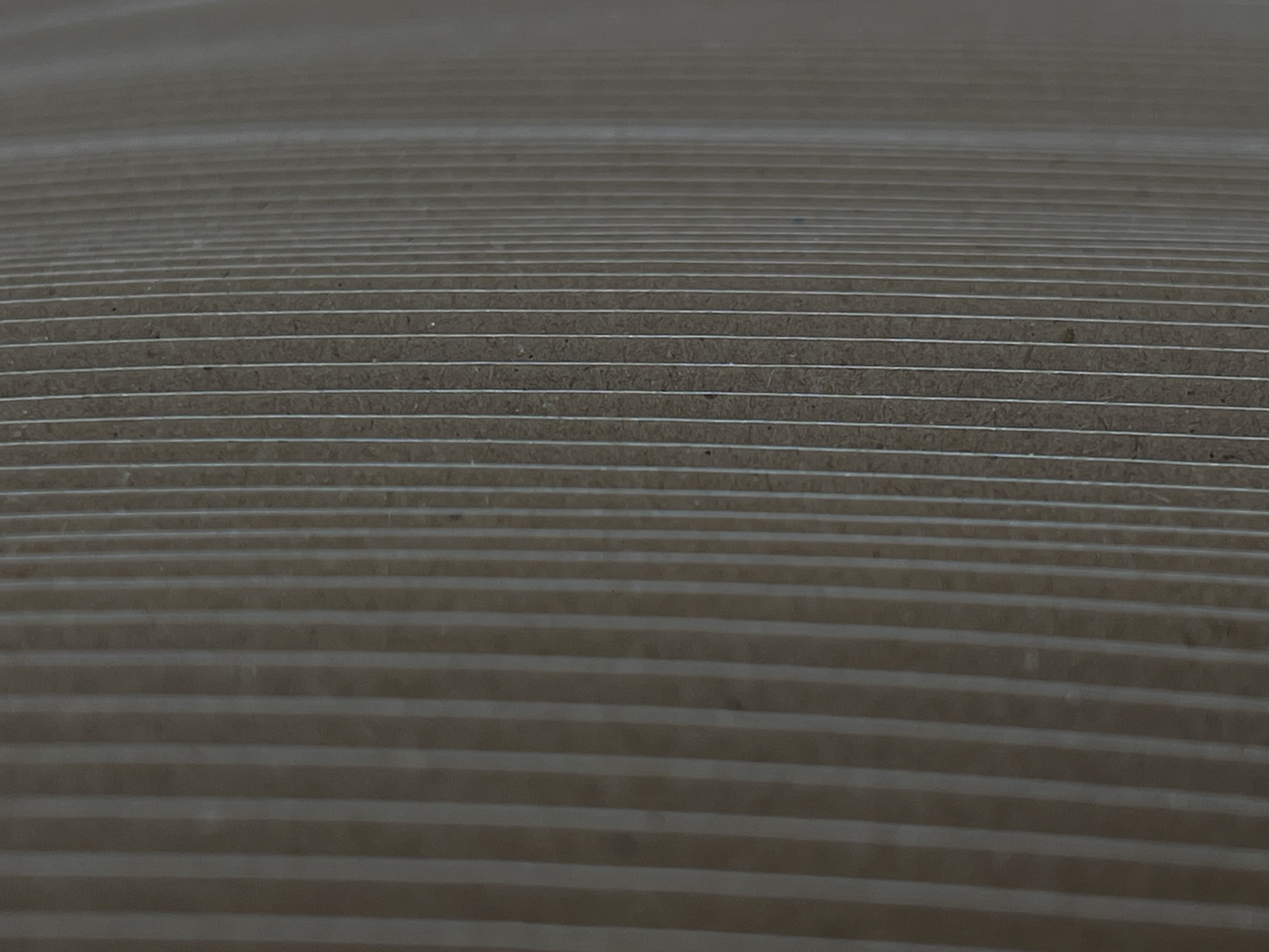 The image depicts a close-up, angled view of stacked, corrugated cardboard sheets. The material is a muted brown color with visible fibrous texture, and thin, lighter-colored lines, likely part of the corrugation, run horizontally across the frame. The perspective suggests the sheets are stacked in a way that creates a visual effect of steps receding into the distance, with the uppermost sheets appearing slightly blurred due to depth of field. The lighting is even and diffused, casting no harsh shadows. There are no people or any identifiable objects other than the cardboard. The scene appears to be an indoor or storage setting, likely related to packaging or manufacturing, and no specific location in Lanžhot, Czechia can be inferred from the visual information provided.