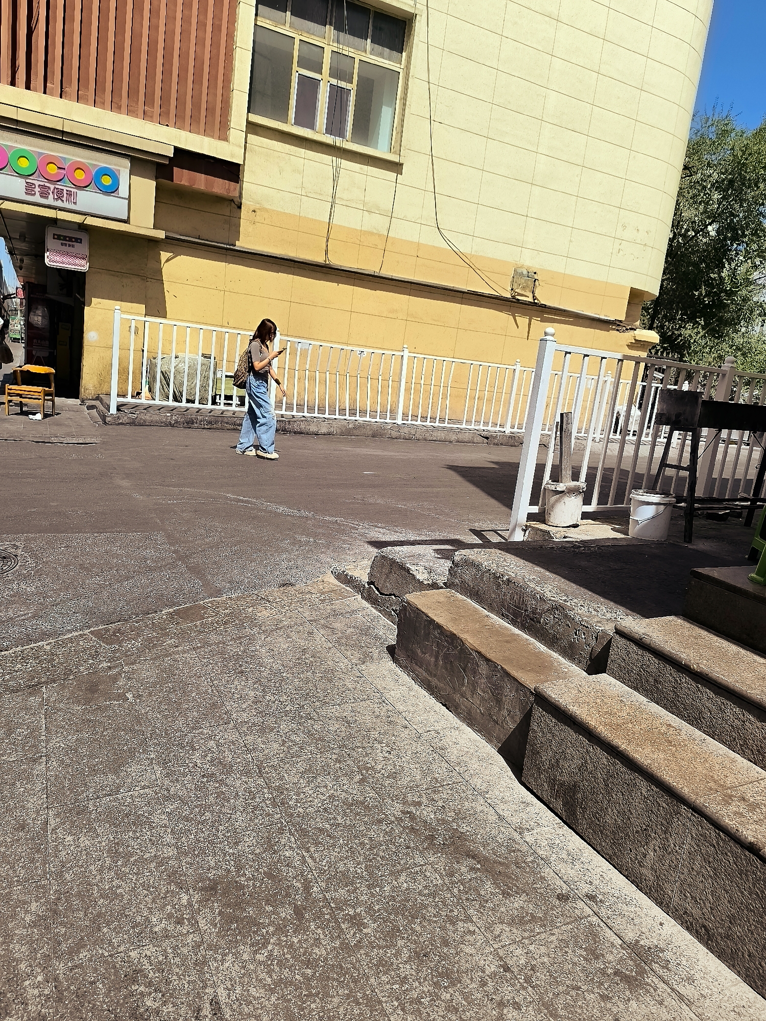 A person walks along a paved street in front of a yellow building. The person is wearing a t-shirt, loose-fitting jeans, and a backpack. The building has a brown corrugated facade on the upper left and a yellow tiled facade on the right. A white metal fence runs along the front of the building. There are two white buckets and a black metal grill behind the fence. In the foreground, there are concrete steps. A sign with colorful circles and Chinese characters is visible on the left side of the building. The scene appears to be in a city, possibly Ürümqi, China, given the sign's characters and the architectural style.