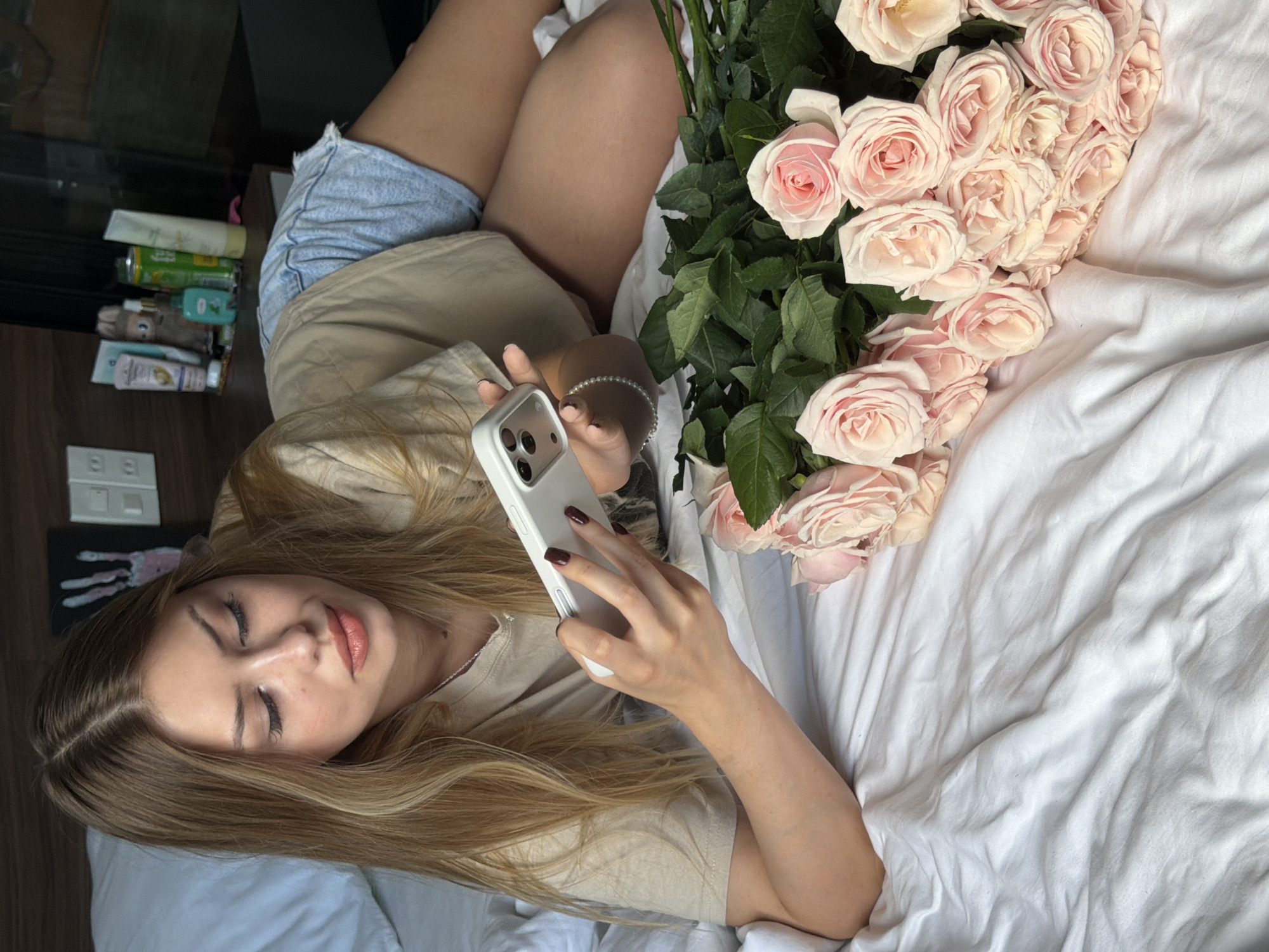 In a bedroom setting, a young Caucasian woman with long blonde hair, wearing a beige t-shirt and denim shorts, is lying on a white bed. She is holding a white smartphone and looking at its screen. To her right, a large bouquet of light pink roses is arranged on the bed. The background shows a wooden headboard with some items on a shelf, including bottles and a framed handprint artwork. The overall scene suggests a private, domestic environment. There are no specific visual cues to indicate the location in the city of Đà Nẵng, Vietnam.
