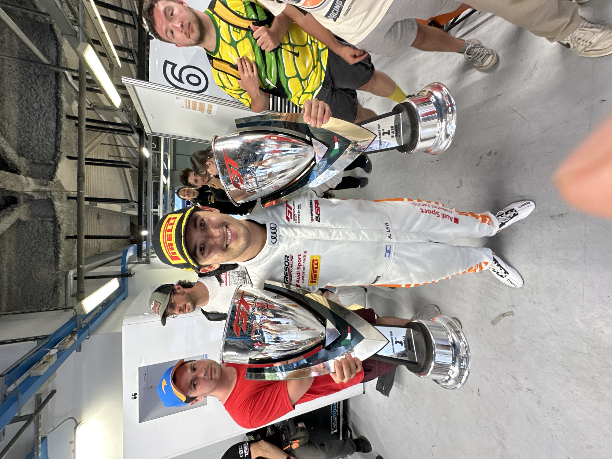 The image captures a celebration indoors, likely in a garage or pit area, with several individuals posing with trophies. In the center, a man in a white and orange racing suit and a yellow Pirelli cap holds one of the trophies. The trophies are silver and display logos such as "WORLD CHALLENGE powered by aws" and "ENDURANCE CUP 1st SILVER Monza", which places the location in Monza, Italy. The suit bears the Audi Sport logo. Other people surround the central figure, including a man in a red shirt and a blue baseball cap holding a trophy, and another man in a yellow and green shirt. The backdrop includes white walls with the number "66" and some overhead industrial structures and lighting. The floor is gray concrete with some markings on it.