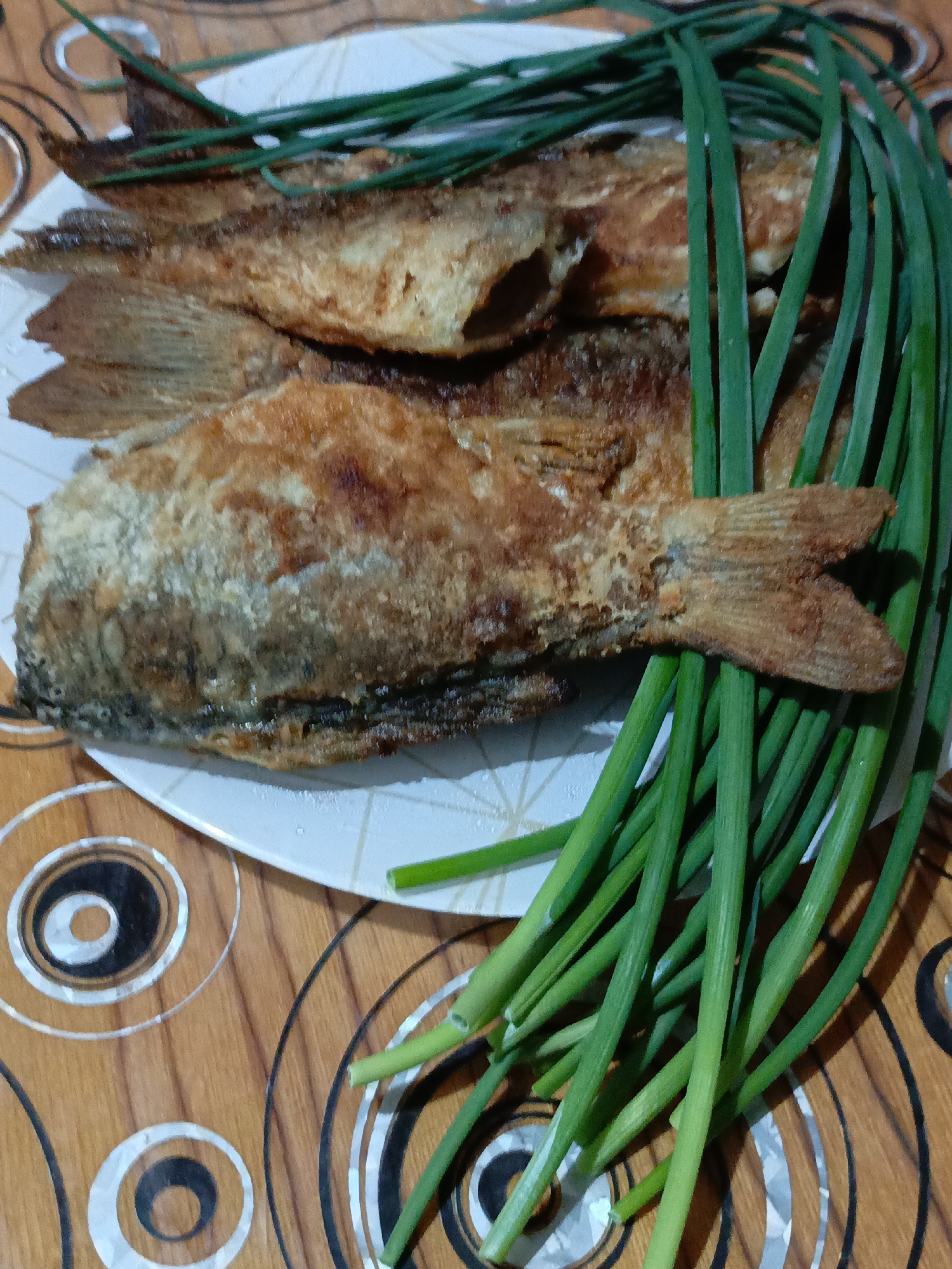 On a patterned, likely wooden surface, a white plate holds three fried fish, each adorned with a fan of fresh green onions. The location is unspecified, though the prompt mentions Zalesovo, Russia.