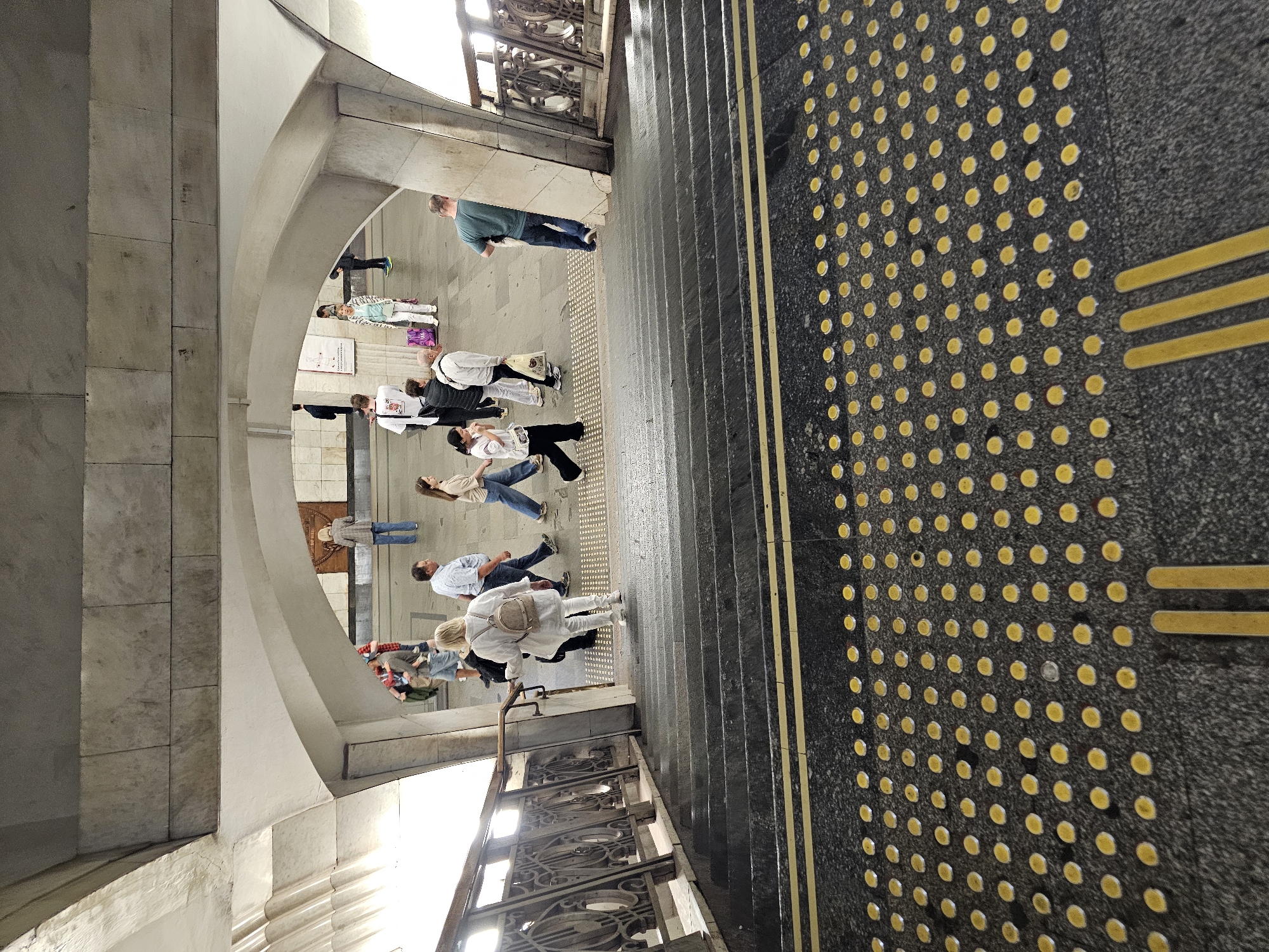 A group of people are seen walking in a subway station in Moscow, Russia. The scene is viewed from a high angle, looking down a wide set of stairs. The stairs are made of dark granite and have yellow tactile paving at the top and bottom, with yellow lines marking the edge of each step. The people are walking in various directions, some are going up the stairs, others are walking on the platform at the top. In the background, there are arches and marble walls typical of a Moscow metro station. The lighting appears to be artificial, as is common in underground stations.