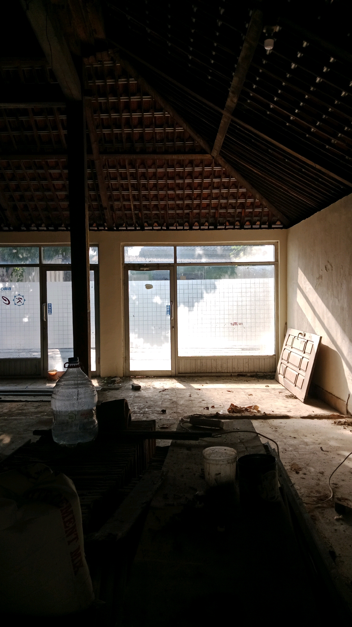 The image depicts the interior of a dimly lit, unfinished or abandoned building, possibly a commercial space, located in Candirejo, Indonesia. Sunlight streams through large glass doors and windows, casting strong diagonal shadows across the dusty, concrete floor. The space is cluttered with various objects, suggesting ongoing or halted construction or renovation.
Prominently in the foreground, a large, clear plastic bottle filled with water stands near a dark, cylindrical object and a large, light-colored bag with visible lettering. Further back, on the floor, are scattered debris, a few pieces of wood, and two containers, one white and one dark. Against the far wall, to the right, rests a wooden door leaning upright.
The architecture of the building is characterized by an exposed, sloped wooden roof with visible rafters and terracotta tiles. Two large glass doors, frosted with a grid pattern, are situated on the left side of the image, while a single large window with a similar frosted pattern spans the majority of the back wall. The walls appear to be rough plaster, showing signs of age and disrepair. The overall atmosphere is one of disuse and a palpable sense of stillness.