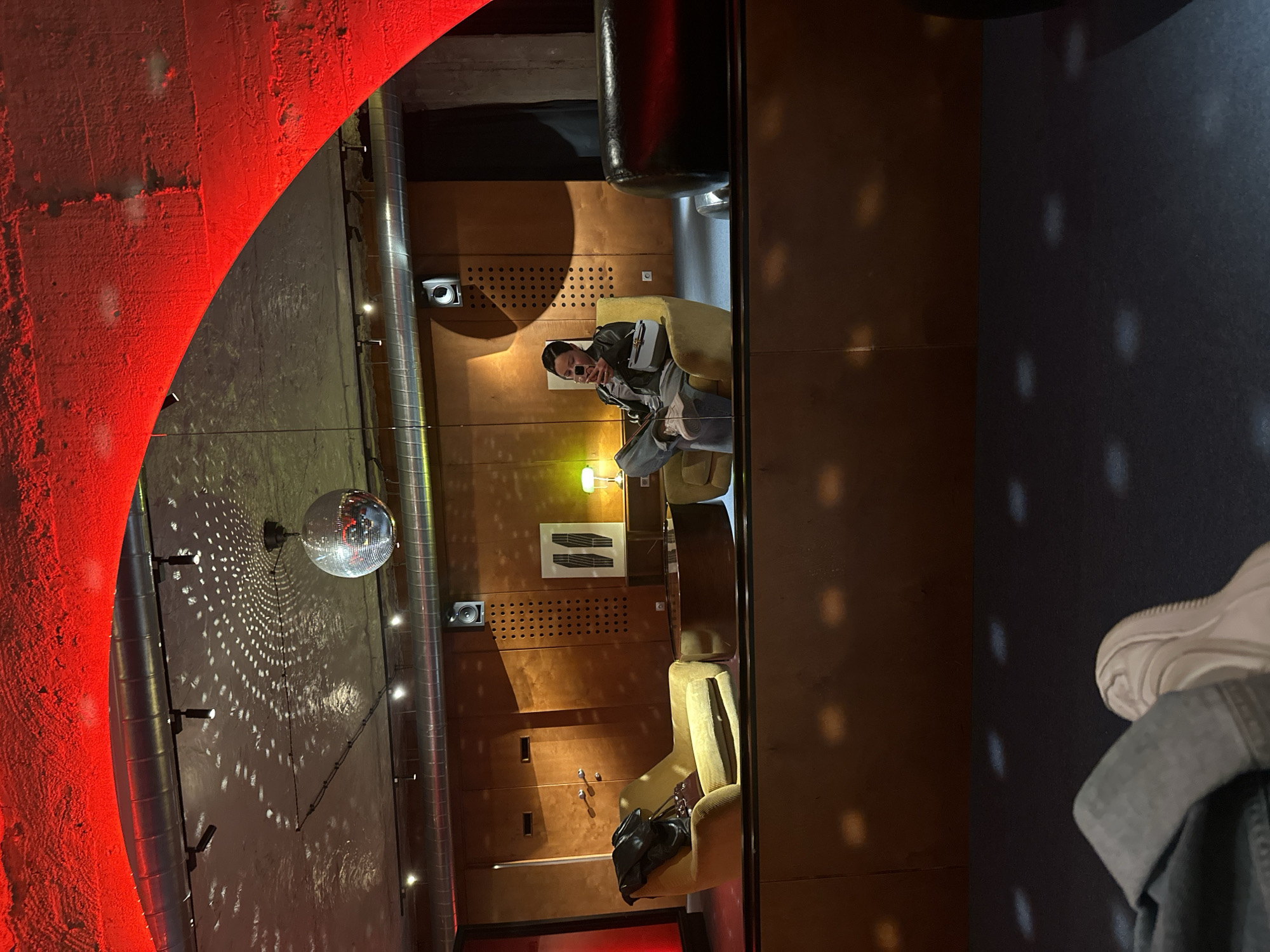 The image depicts the interior of a bar or lounge in Yerevan, Armenia. The room features a disco ball hanging from the ceiling, casting light patterns on the walls. A person is seated in a yellow armchair against a paneled wall with speakers and decorative elements. A second person is partially visible in the foreground. The room is bathed in red lighting.