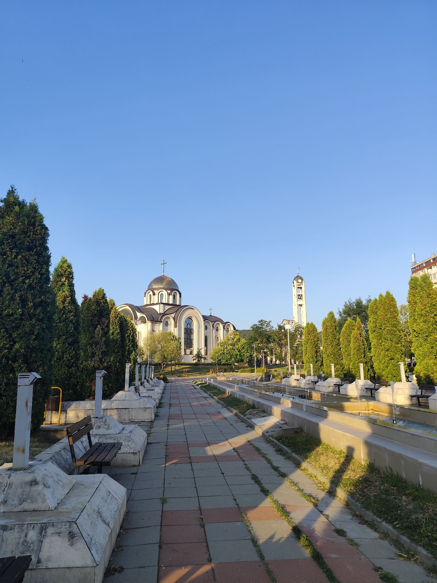 The image depicts a view of the "St. Cyril and Methodius" Orthodox Cathedral in Montana, Bulgaria. The cathedral, with its golden dome and white facade, is centrally located in the frame. In front of the cathedral, a pathway paved with rectangular tiles in shades of gray and red leads towards it. Flanking the pathway are rows of trees, mostly cypress-like in shape, and a series of modern fountains with water jets. Benches are visible behind the decorative stone structures lining the fountains. To the right of the cathedral, a tall bell tower stands against a clear blue sky. In the background, a multi-story building can be seen. The scene is bathed in the warm light of what appears to be late afternoon or early morning, casting shadows from the trees and pathway onto the grass. The overall impression is one of a well-maintained public space or park adjacent to a prominent religious building.