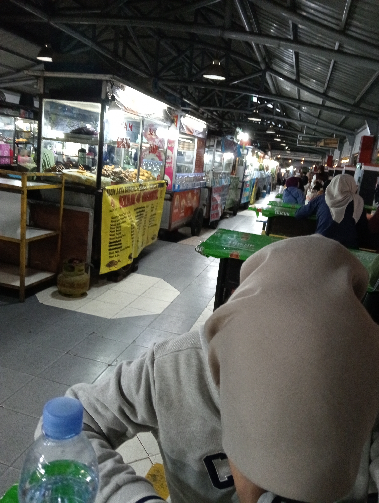 This is a medium shot of a covered person sitting at a food court in Magelang, Indonesia. The person is wearing a light-colored head covering and a grey sweatshirt. In the foreground, a partially filled plastic water bottle with a blue cap is visible. The background shows a row of food stalls with various items displayed, including fried foods and drinks. Several other people are seated at tables, some with head coverings. The food court appears to be covered by a metal roof structure with visible beams and lighting fixtures. The floor is tiled with light-colored octagonal and square tiles. A yellow banner with text is displayed at one of the stalls. A gas cylinder is visible near a shelf unit. The overall atmosphere suggests a casual dining environment.