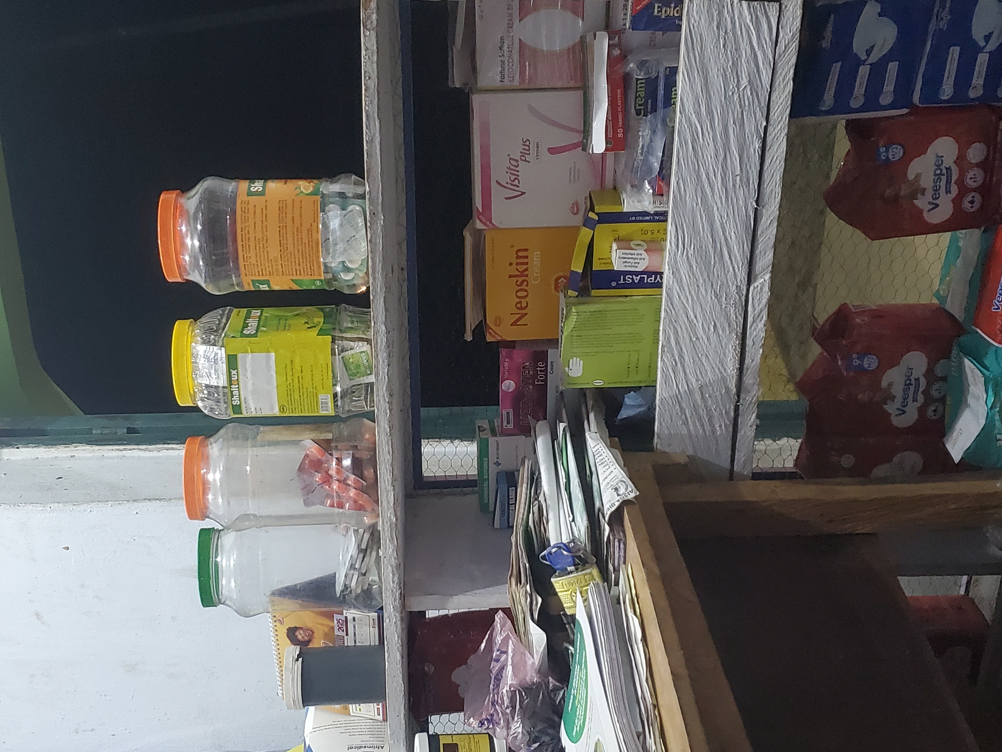 This is a medium shot, eye-level photograph taken in a retail setting, likely a pharmacy or small shop, in Mutum Biyu, Nigeria. The image depicts a cluttered shelf filled with various pharmaceutical products and supplies.
In the background, three large, clear plastic jars with colorful lids (green, orange, and yellow) contain what appear to be pills or capsules. Labels on these jars indicate brands like "Shaltlux." To the left of the jars, a smaller dark grey container sits next to a spiral-bound calendar showing the year 2025 with a picture of a smiling person.
The middle shelf is crowded with stacked items. A collection of papers and documents is piled on the left side, some held together by a blue lock and key. Various boxes of medical supplies are visible, including "Neoskin Cream," "Visita Plus Cream," and "Fortune Saffron Ketoconazole Cream BP." There are also smaller boxes labeled "TYPLAST" and others containing fabric plasters and surgical blades.
The lower shelf displays larger packages of what appear to be sanitary products or diapers, branded "Veesper." Boxes of other items, possibly pharmaceutical or hygiene products, are stacked to the right. The shelves themselves appear to be made of wood and possibly metal, with a textured white surface. The overall environment suggests a busy, functional space where goods are stored and displayed for sale. The lighting is somewhat dim, contributing to a slightly shadowy atmosphere.
