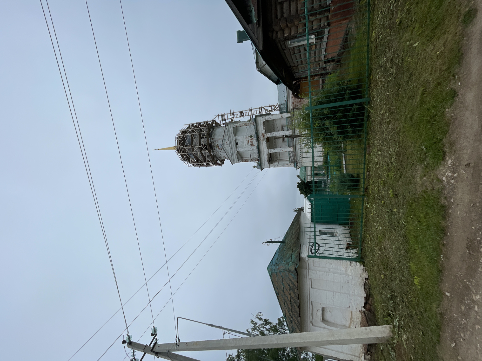The image shows a view of a church bell tower under scaffolding, with surrounding buildings and infrastructure in Кстинино, Russia. A white, multi-story bell tower with classical architectural elements, including columns and arched windows, stands prominently in the center-right of the frame. The upper portion of the tower is covered in a wooden scaffolding structure, suggesting ongoing construction or restoration. A golden spire tops the bell tower. To the left of the bell tower is a small, white, single-story building with a green, weathered, sloped roof. A green metal fence encloses the area in front of this building and extends towards the bell tower. To the right of the bell tower, a portion of a dark, wooden log house is visible. Overhead, power lines crisscross the cloudy, gray sky, supported by a concrete utility pole on the left side of the image. The ground in the foreground is unpaved, with a dirt and gravel surface transitioning into a strip of green grass.