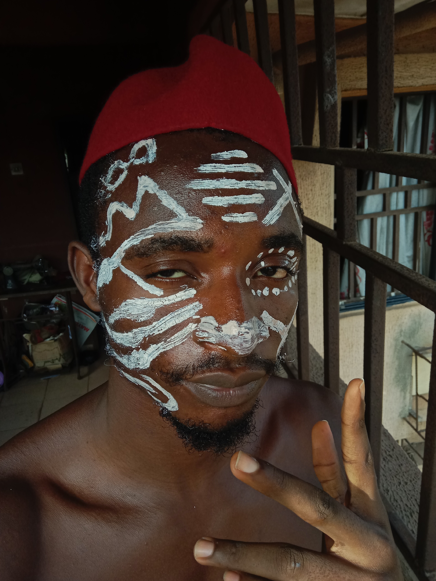 A close-up of a man with dark skin. He is wearing a red cap, and his face is painted with white markings. He is standing in front of a metal railing. He is holding up his hand in a peace sign. The location is Nkpor, Nigeria.