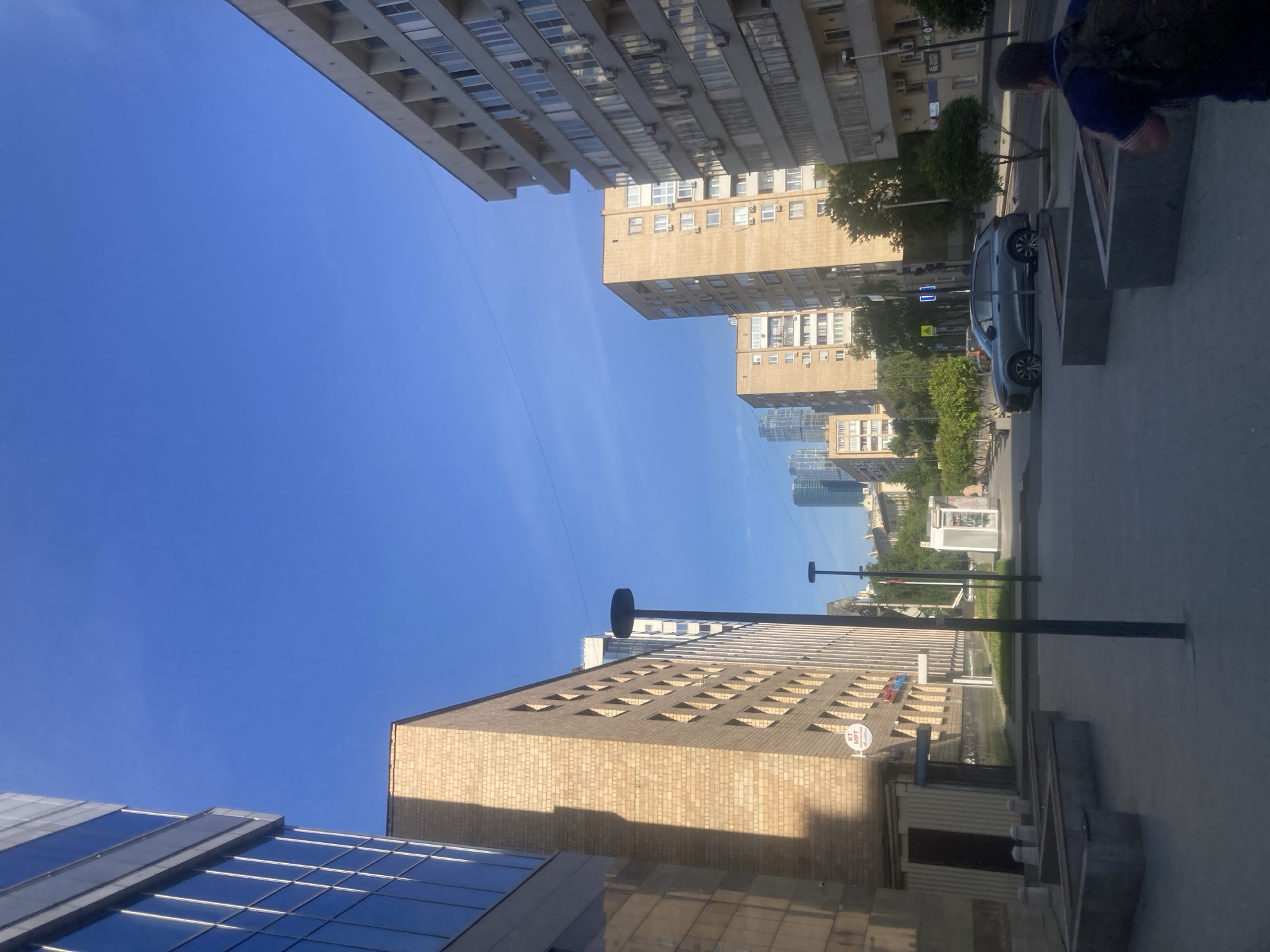 The image depicts a street scene in Moscow, Russia, with tall buildings lining the roadway. The architecture of the buildings varies, with some constructed from brick and others from glass and steel. The sky is blue and mostly clear. A car is parked along the street, and a person stands near it. Streetlights are visible along the sidewalk. The scene appears to be taken during the daytime with good lighting.