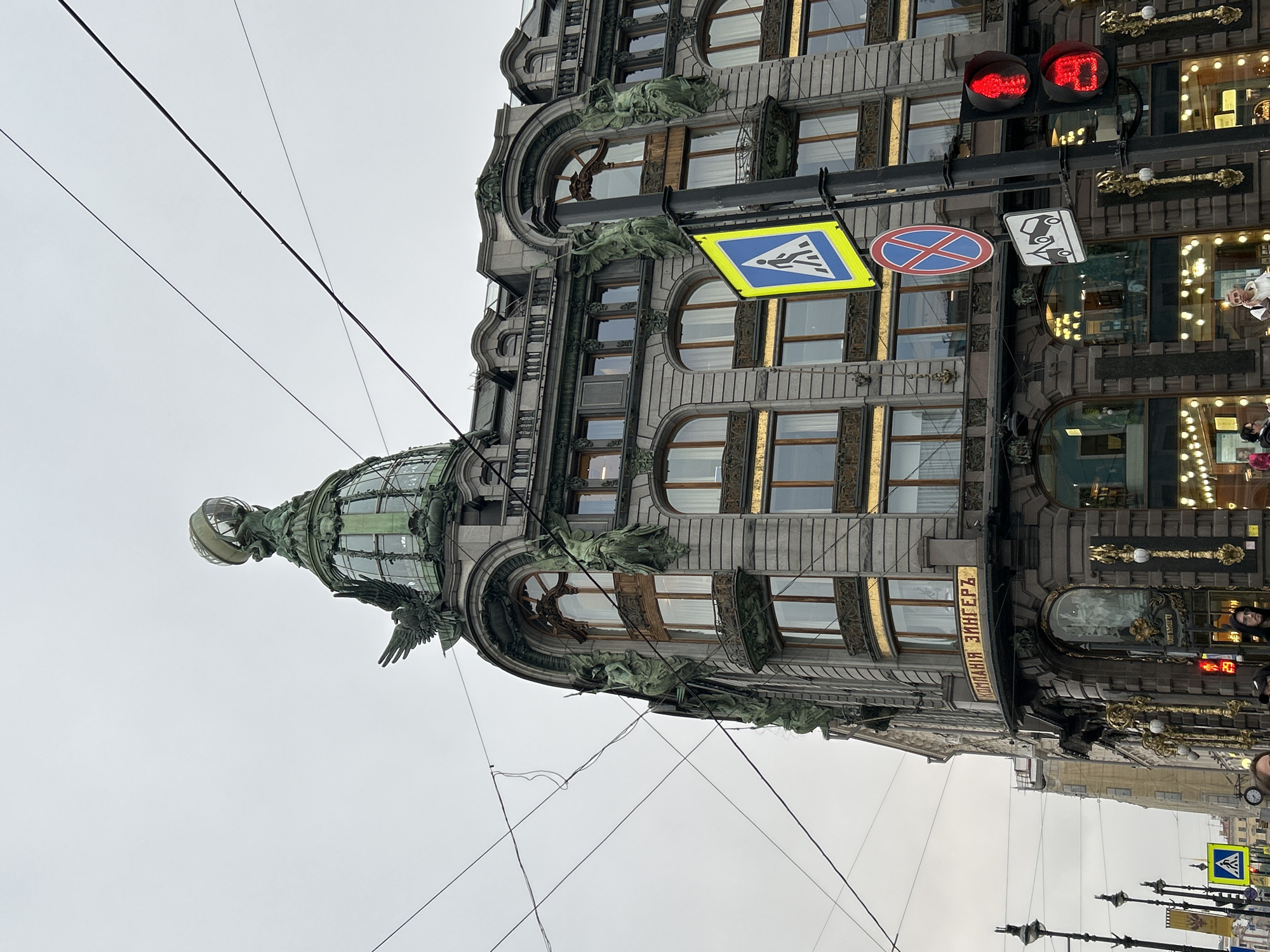 This is a photograph of the Singer House building, also known as the House of Books, located in Saint Petersburg, Russia. The ornate building features architectural details such as arched windows, decorative sculptures, and a glass dome topped with a sculptural globe. The building appears to have multiple stories. Utility lines are visible stretching across the sky in the foreground. Street signs and traffic signals are mounted on a pole attached to the building. The signs include a pedestrian crossing symbol and a "no parking" sign. A red traffic light is visible. The building's name, "КОМПАНIЯ ЗИНГЕРЪ" (Company Singer), is inscribed on the facade. The sky is overcast, suggesting a cloudy day. Pedestrians and other buildings are visible in the background, indicating an urban setting.