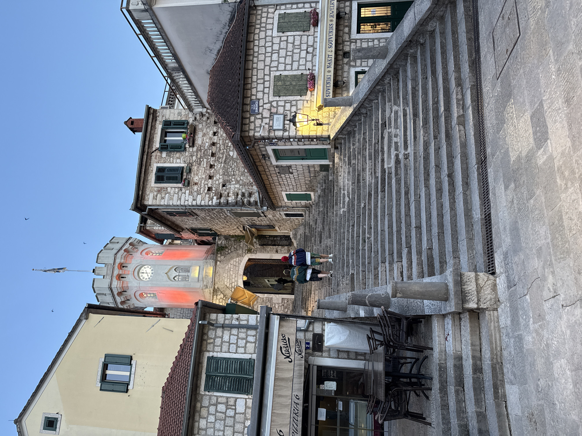 Here is a description of the image:
The image depicts a street scene in Herceg Novi, Montenegro, featuring its notable clock tower. The tower has a cream-colored facade with red lighting highlighting the architectural details. A clock face is visible near the top. The surrounding buildings have stone facades with green shutters on the windows. Two people walk up a set of wide stone steps. A restaurant or pizzeria named "Musica" with outdoor seating is in the foreground. The sky is a light blue color.