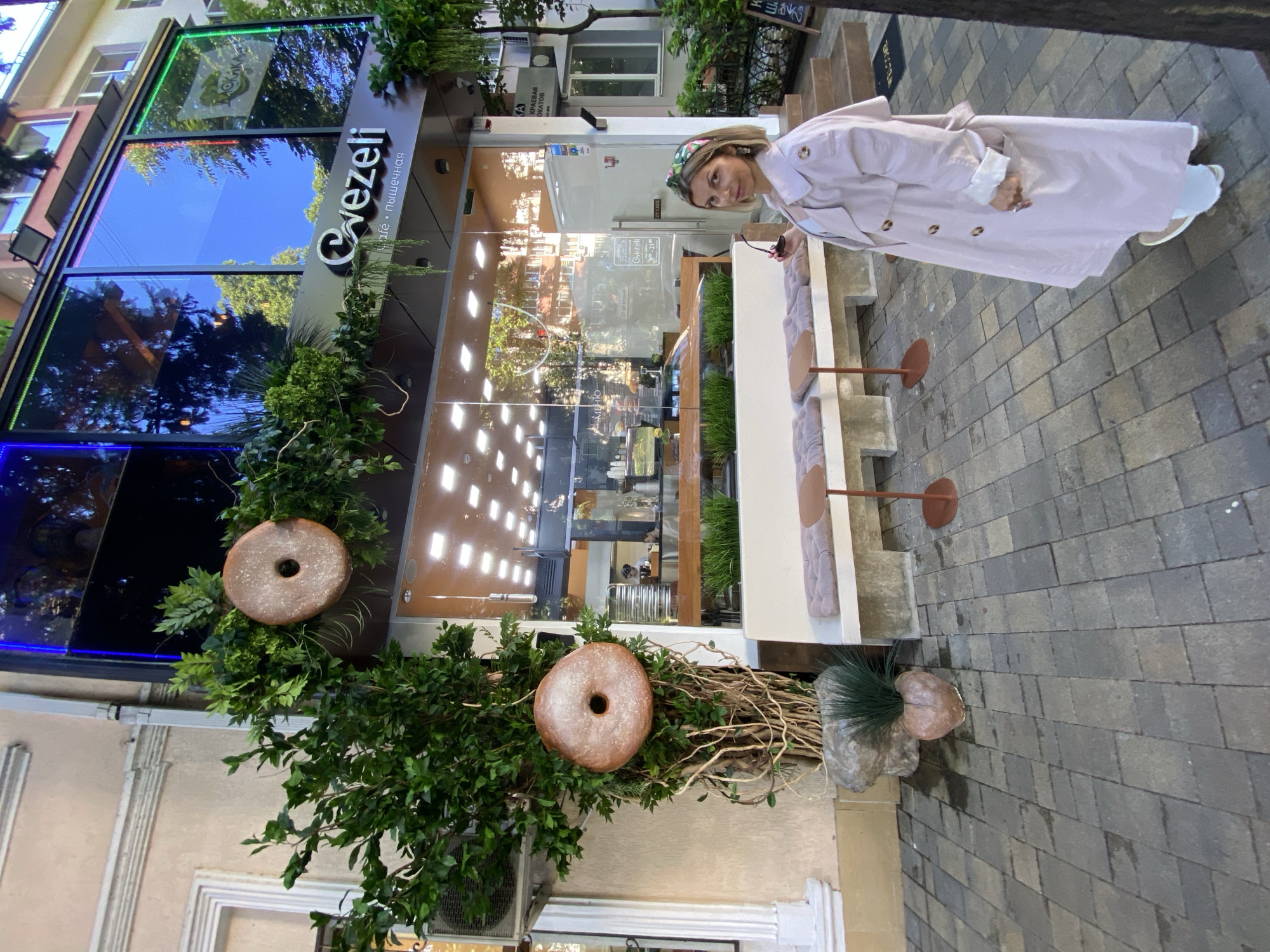 An image captures a young woman posing in front of a modern storefront in Sochi, Russia. She stands beside an outdoor seating area, leaning on a long, white bench with light-colored cushions. The woman wears a light-colored (possibly pale pink or beige) long trench coat over white wide-leg trousers and light flat shoes. She has a patterned headband and earrings, looking towards the camera with a slight smile.
The storefront features large glass windows that reflect the blue sky, green trees, and other buildings, creating a bright, open feel. Above the entrance, a sign prominently displays the stylized name "Cvezeli" (or "Gvezeli") followed by the Cyrillic text "кафе • пышечная," which translates to "cafe • donut shop" or "cafe • pyshka shop" (pyshka being a type of Russian donut). The facade is extensively decorated with artificial greenery and branches, from which two large, donut-shaped sculptures protrude, reinforcing the shop's theme.
In front of the bench, there are two small, round, brown tables. The ground is paved with grey and dark grey rectangular tiles. An air conditioning unit is visible on a building in the background, and a "WELCOME" mat is partially seen near an entrance. The scene takes place during the daytime under sunny conditions, suggesting a pleasant urban setting.