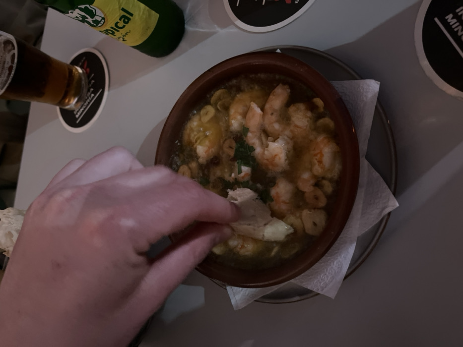 A hand is dipping a piece of bread into a bowl of shrimp and garlic in a clay bowl, sitting on a white napkin on a gray table. The bowl sits on a gray plate. The hand is out of focus, and the table is also out of focus, but the bowl and the food are in focus. The bowl is full of shrimp and garlic, and there is some parsley sprinkled on top. There is a glass of beer and a bottle of beer in the background, out of focus. The beer bottle is green, and the beer is amber. The location is in a restaurant in Arucas, Spain, based on coordinates 28.118 latitude, -15.524 longitude.