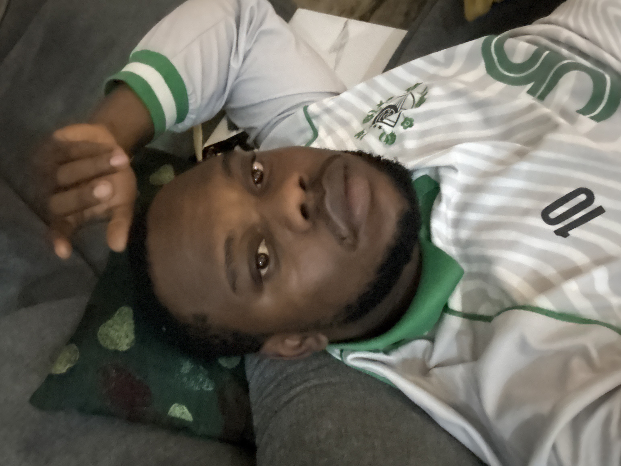 A man is lying down on a grey couch, resting his head on a dark green patterned pillow. He is wearing a grey and white striped jersey with the number 10 on it. The jersey has green accents on the collar and around the sleeves. The location is unspecified but inferred to be Iddo Tudunwada, Nigeria.