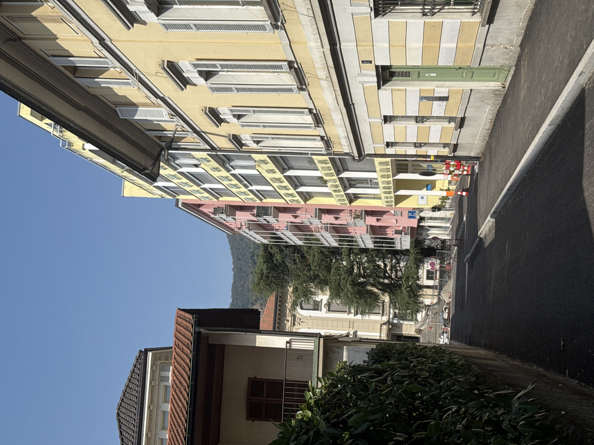 The image depicts a street scene in Chiasso, Switzerland, under a clear blue sky. The perspective is from a street-level viewpoint looking down a relatively narrow road flanked by buildings.
On the left side of the street, a wall covered in dense green foliage runs along the foreground. Behind the foliage, a building with a wooden balcony and shuttered windows is visible. Further down the street on the left, another building with a red tiled roof and decorative architectural elements is partially obscured by trees.
The right side of the street is dominated by buildings with distinct architectural styles. Closest to the foreground is a building with a yellow facade featuring horizontal stripes of white and yellow. This building has numerous windows with white shutters and ornate ironwork grates. A prominent green door is visible on this building, as well as a small sign with the number "2". Further down the street on the right, a tall, slender building with a pink facade and balconies is visible. This is followed by a yellow building with a repetitive pattern of windows and decorative elements.
The street itself appears to be newly paved or undergoing resurfacing, with cones and barriers indicating construction work. The road surface is dark asphalt, and a new concrete curb runs along the right side of the street. In the distance, lush green trees and hills are visible, suggesting a suburban or semi-urban setting with proximity to nature. The overall impression is one of a well-maintained street in a town with a mix of historical and modern architecture.